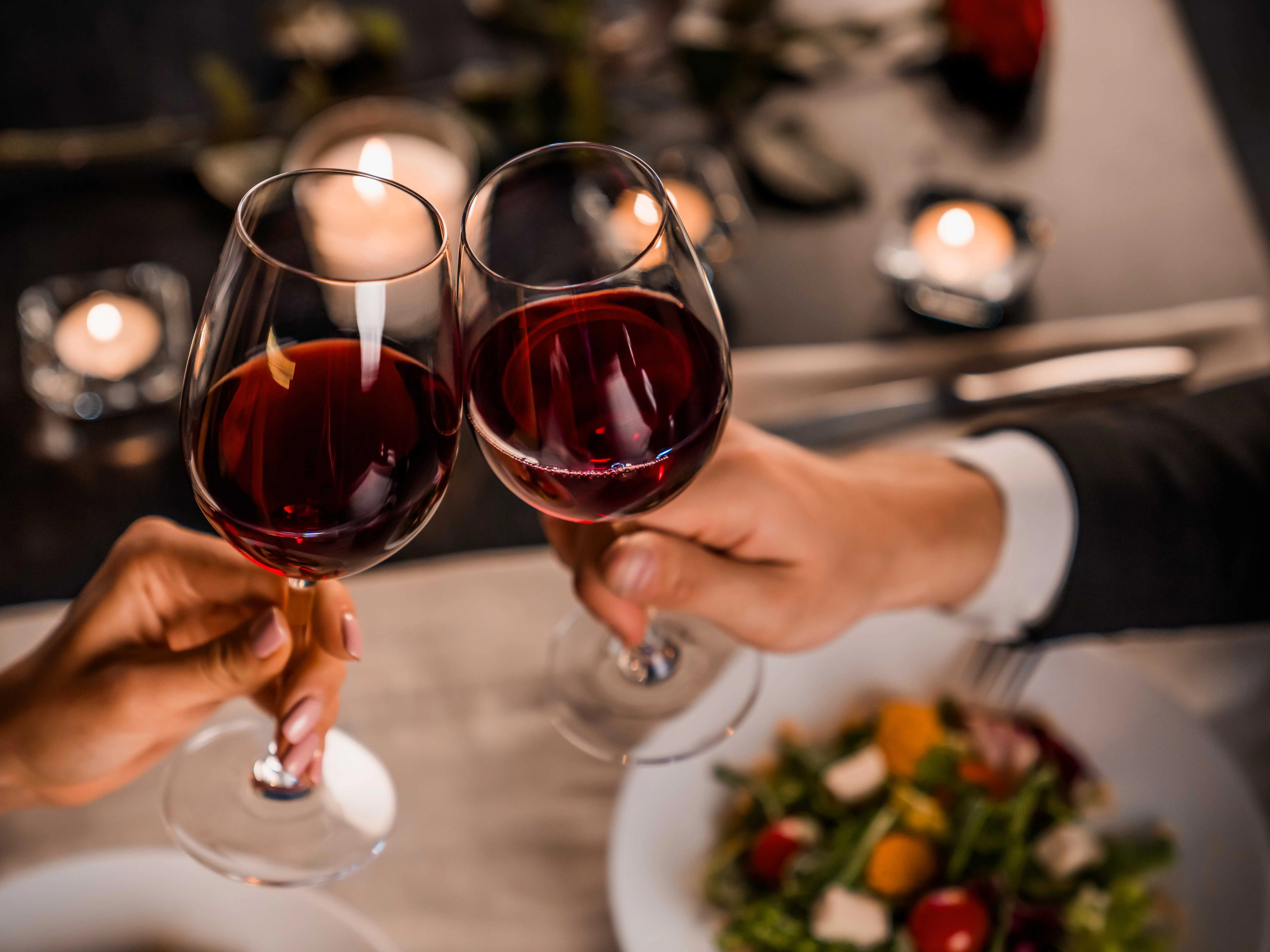 Two people clinking glasses of red wine over a candlelit dinner table.