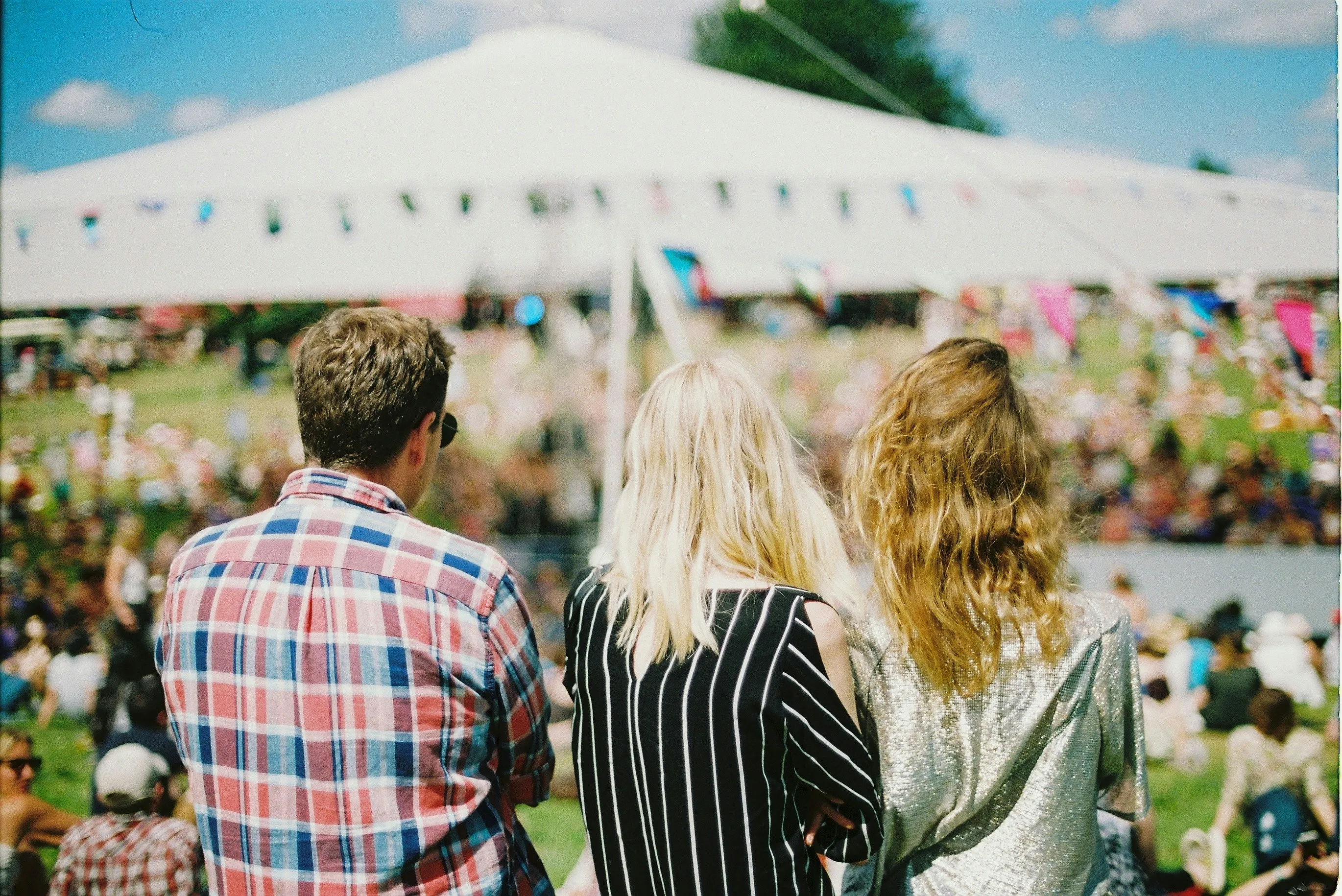 Three people seen from behind at an outdoor festival facing a large white tent and crowd.