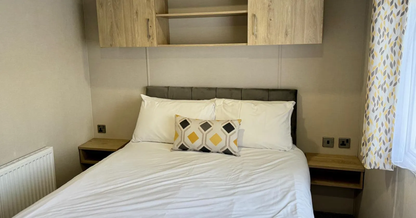Double bedroom with overhead storage cupboards, bedside shelves and patterned curtains in the Carnaby holiday home