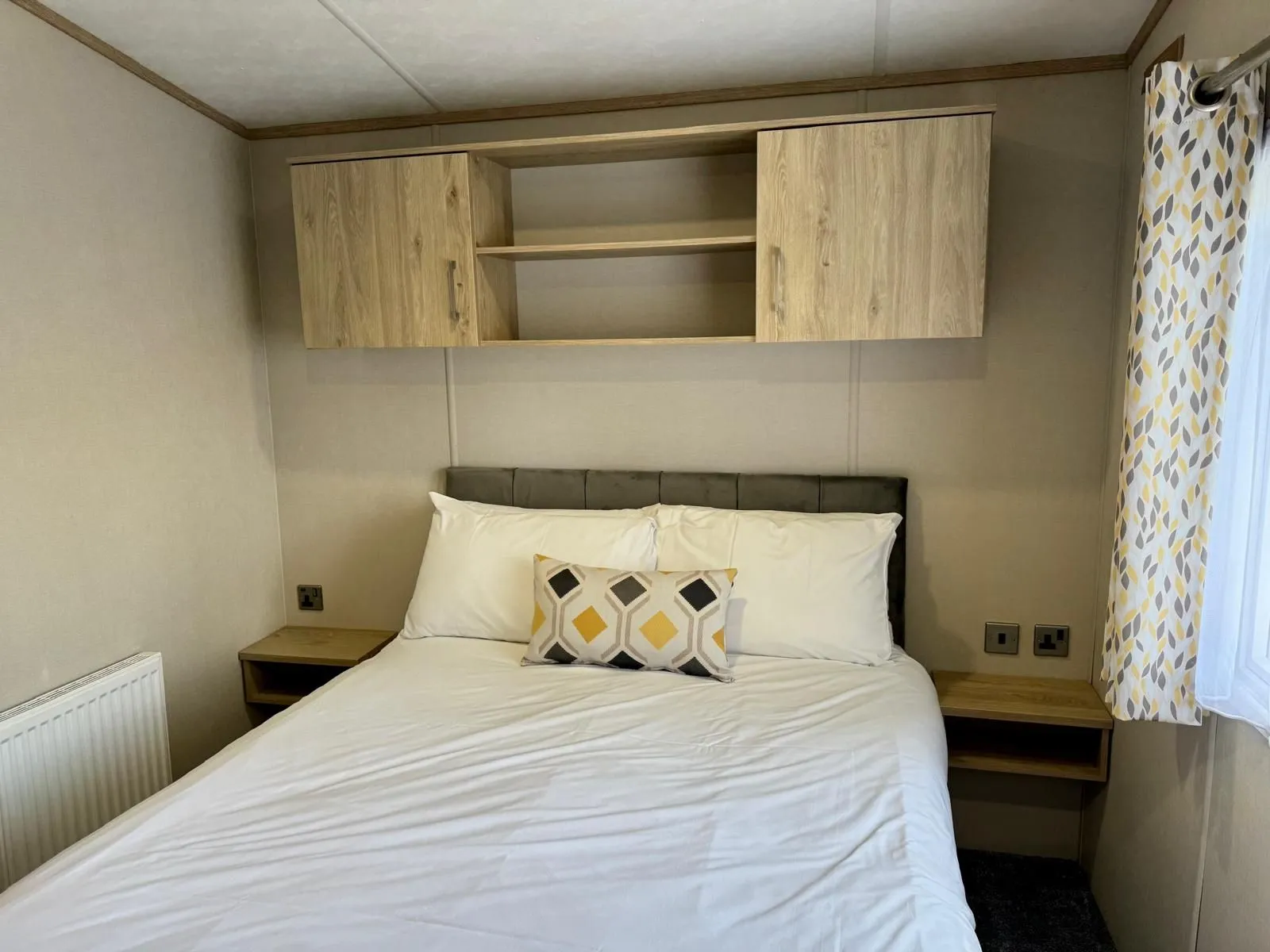 Double bedroom with overhead storage cupboards, bedside shelves and patterned curtains in the Carnaby holiday home