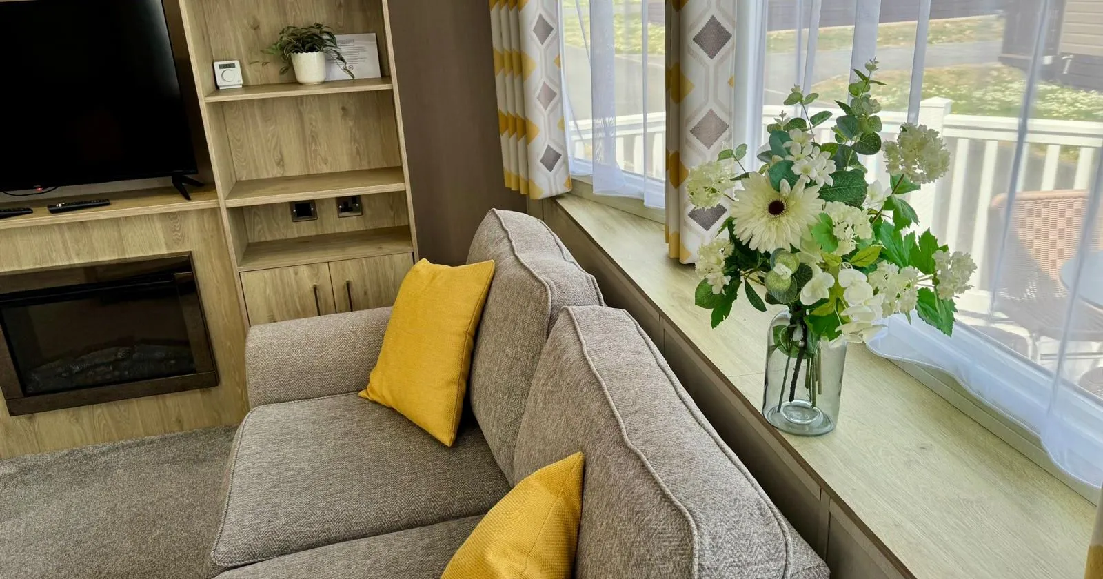 Modern caravan lounge with a grey sofa, yellow cushions, floral arrangement, and TV unit by a large window