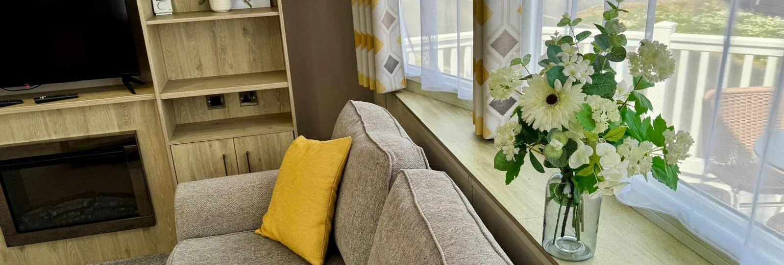 Modern caravan lounge with a grey sofa, yellow cushions, floral arrangement, and TV unit by a large window
