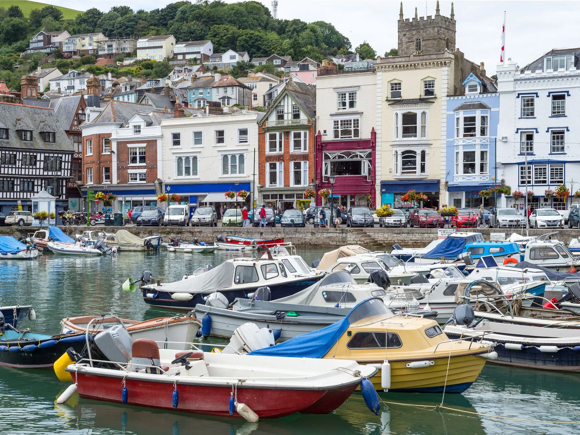 Boats moored in a colourful harbour with waterfront buildings and a church tower on a hillside town