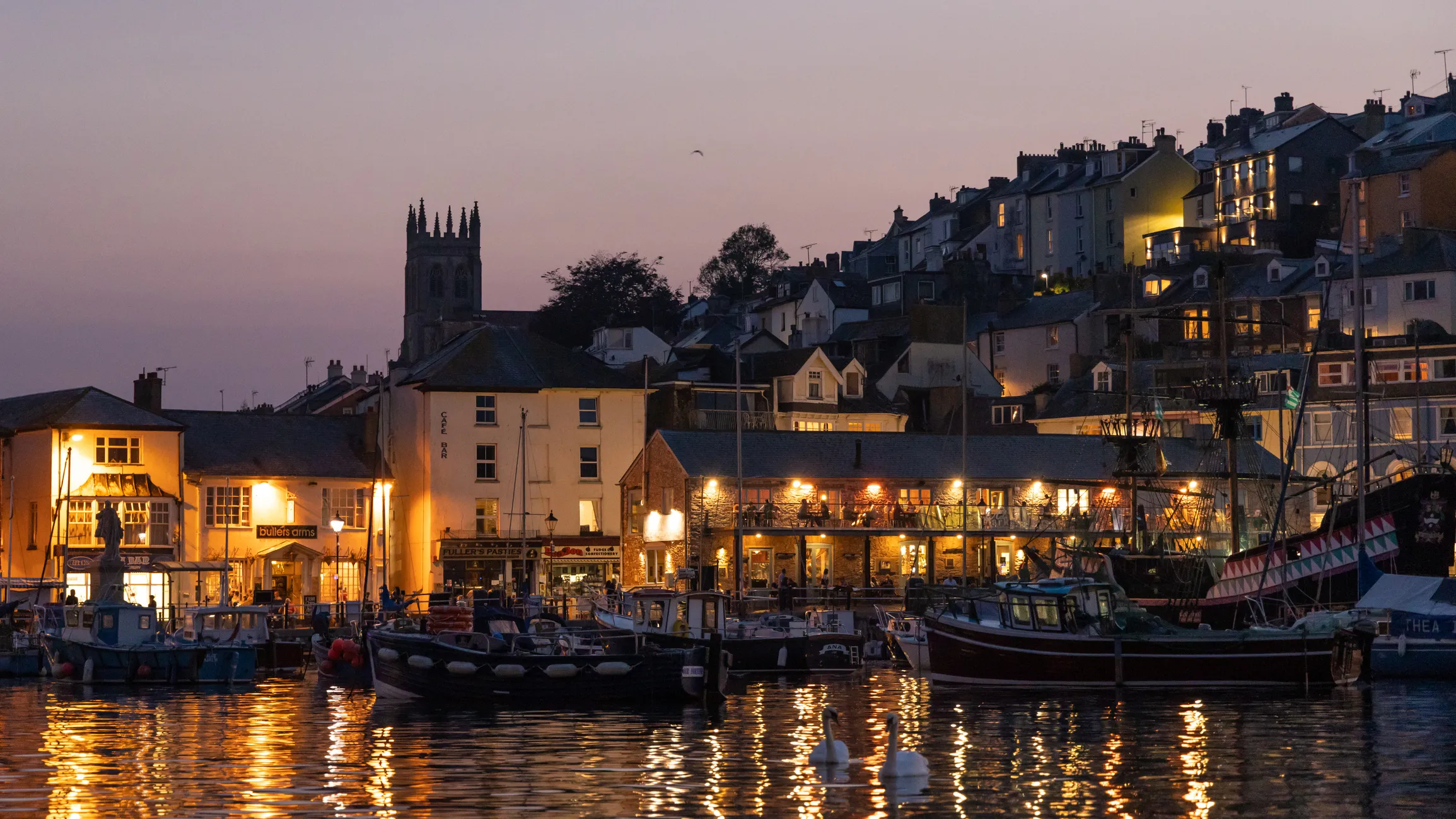 Harbour at dusk with fishing boats, glowing waterfront buildings and hillside houses reflected in the water