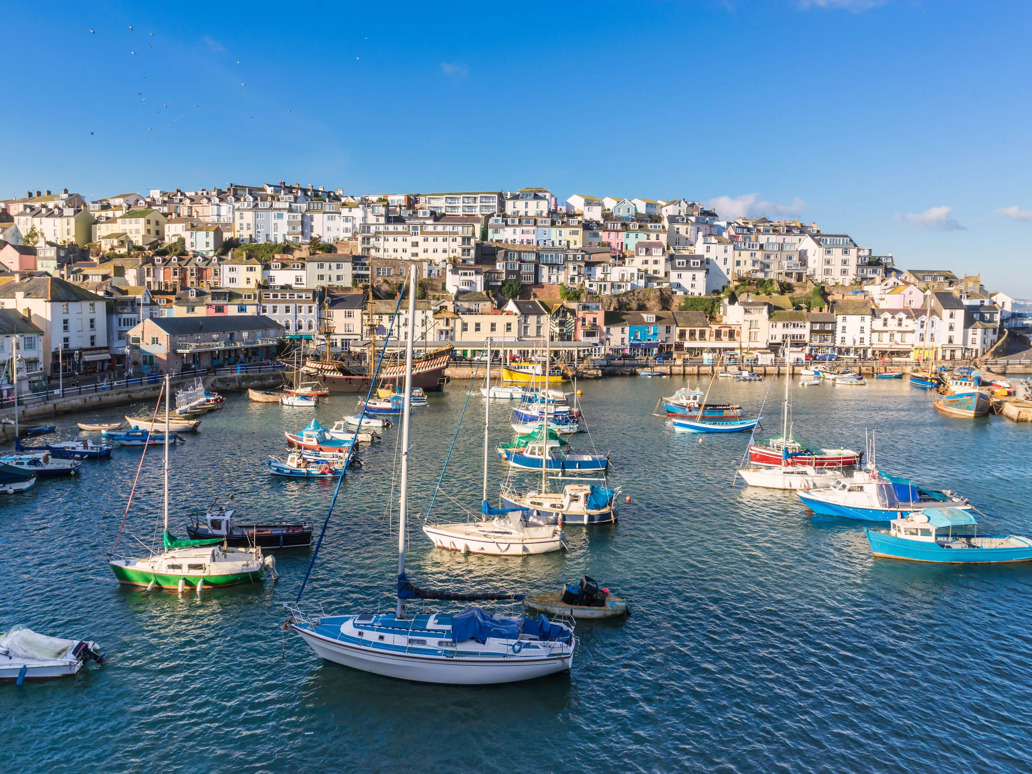 Colourful hillside houses overlooking a harbour filled with small sailing boats and fishing vessels under a clear blue sky.