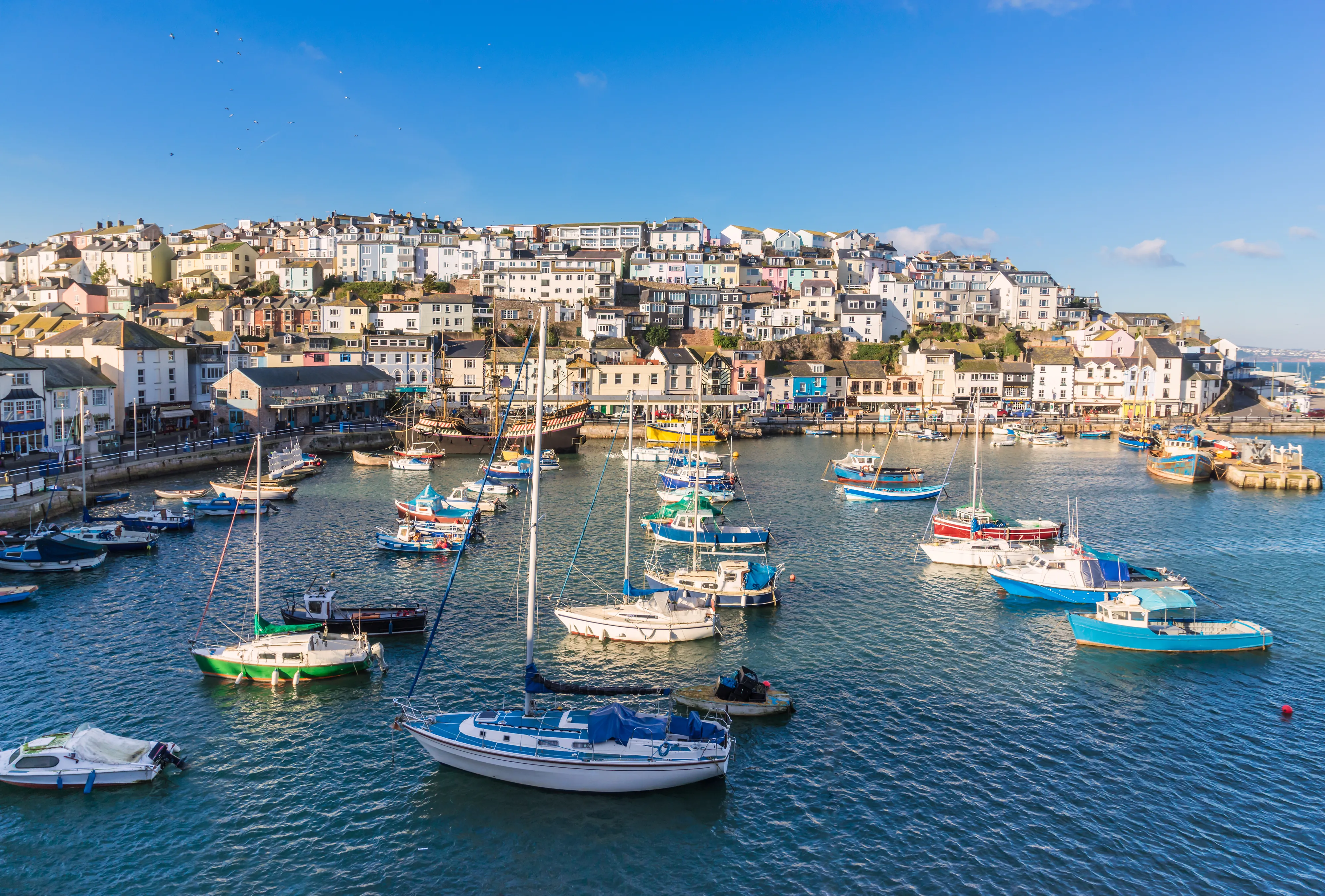 Colourful hillside houses overlooking a harbour filled with small sailing boats and fishing vessels under a clear blue sky.