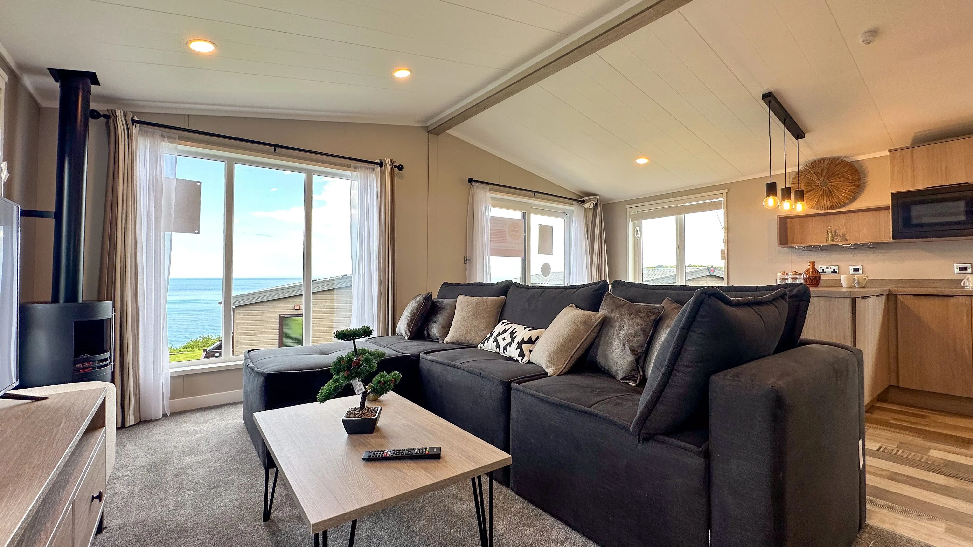 Modern open-plan living room with a large dark sofa, wood-burning stove and sea view through wide windows