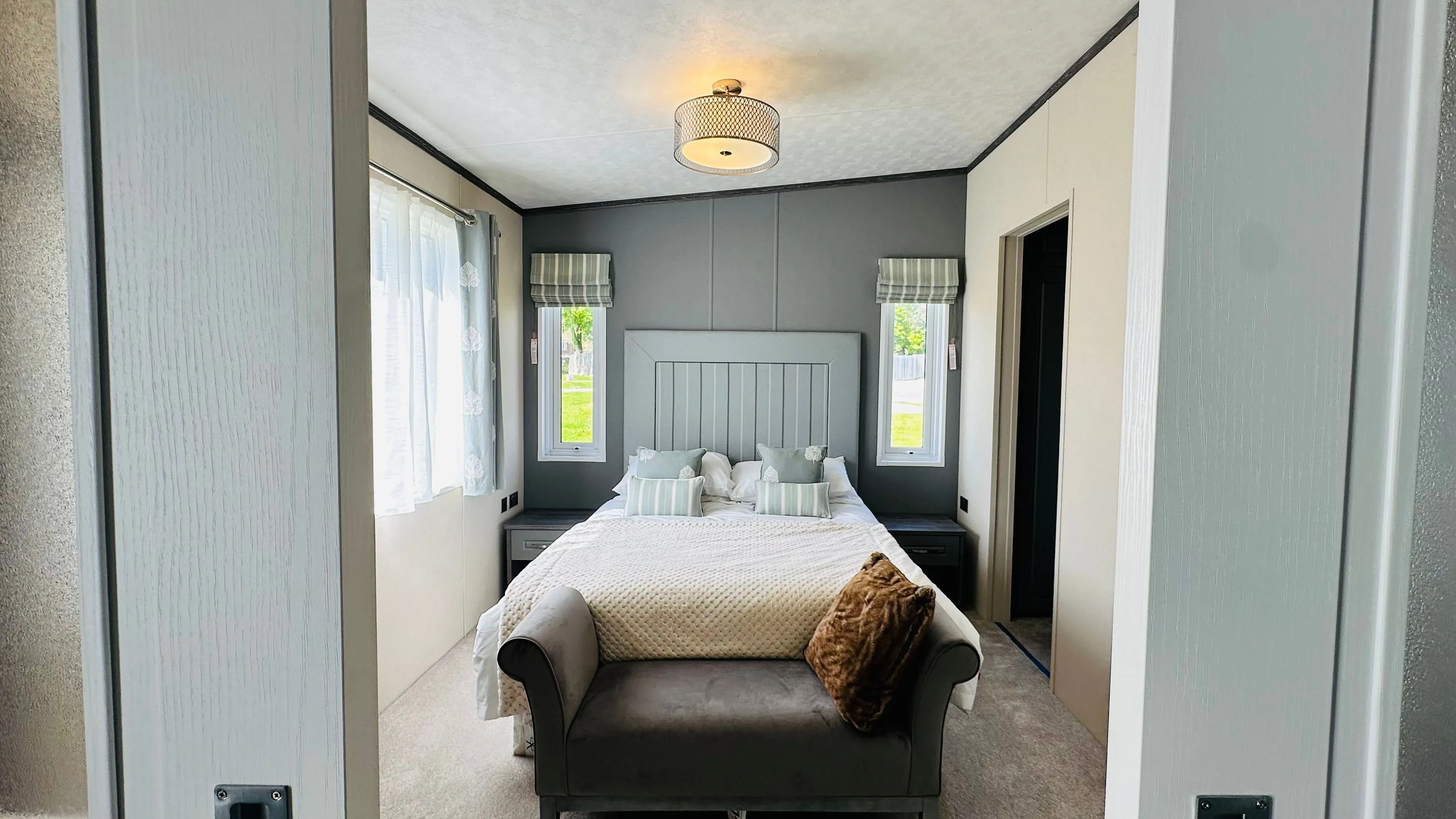 Modern bedroom viewed through sliding doors, featuring a double bed, grey accent wall, bedside tables and a cushioned bench at the foot of the bed.