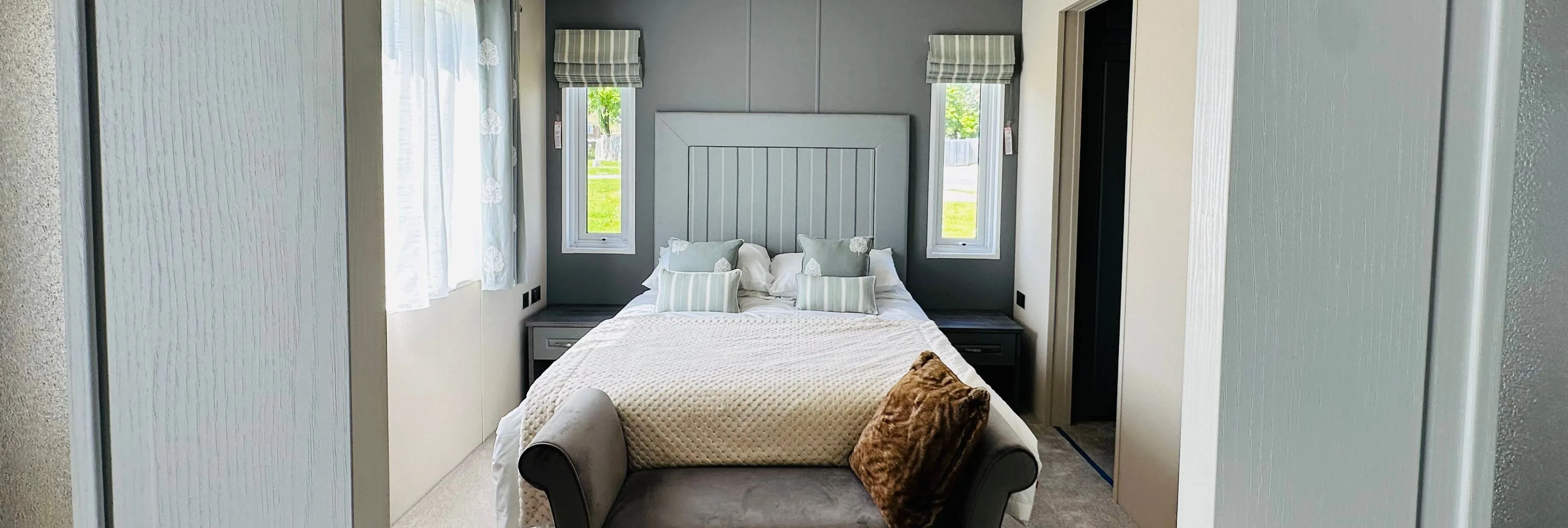 Modern bedroom viewed through sliding doors, featuring a double bed, grey accent wall, bedside tables and a cushioned bench at the foot of the bed.