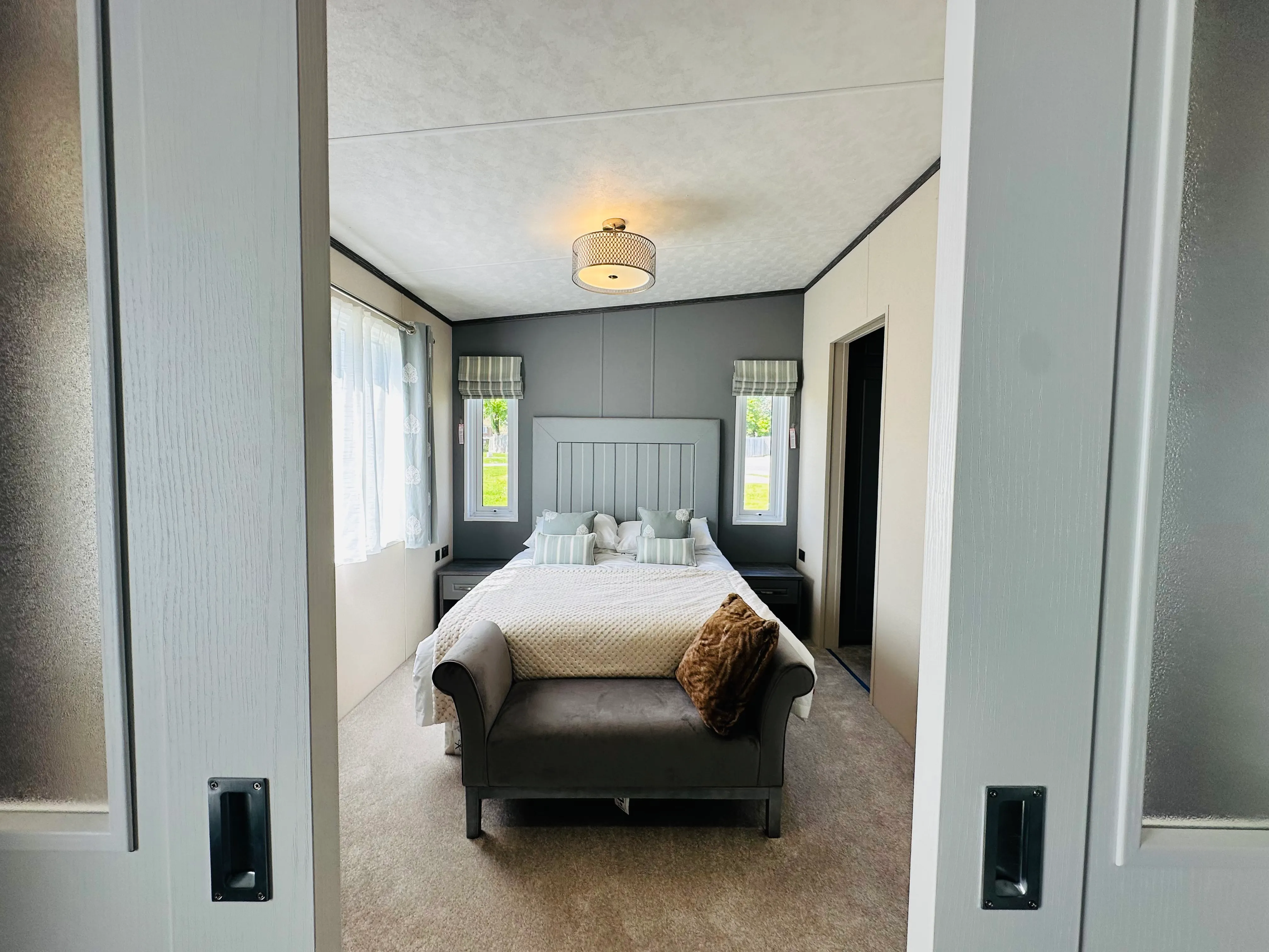 Modern bedroom viewed through sliding doors, featuring a double bed, grey accent wall, bedside tables and a cushioned bench at the foot of the bed.
