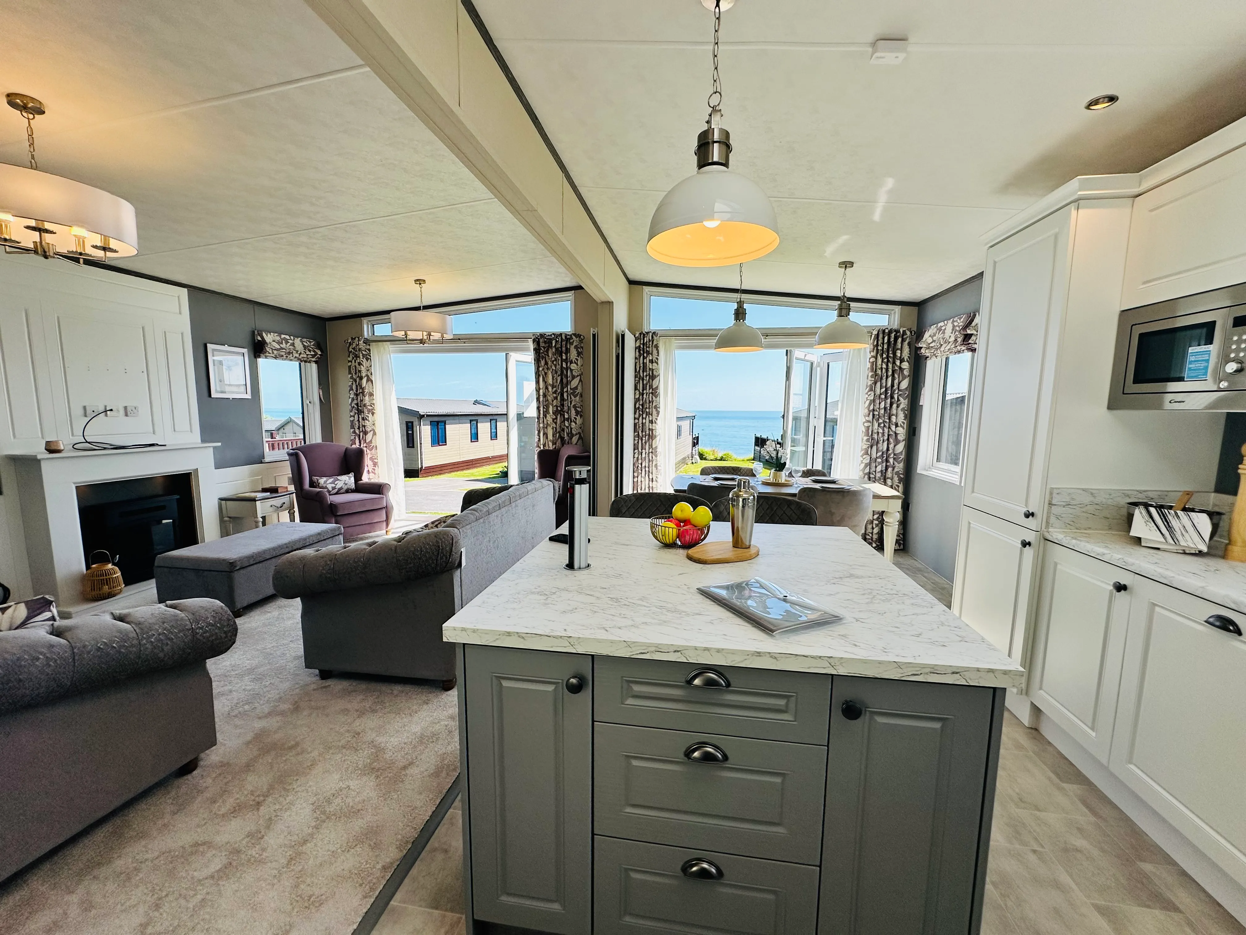 Modern open-plan holiday lodge interior with kitchen island, lounge area, dining space and sea view through large windows