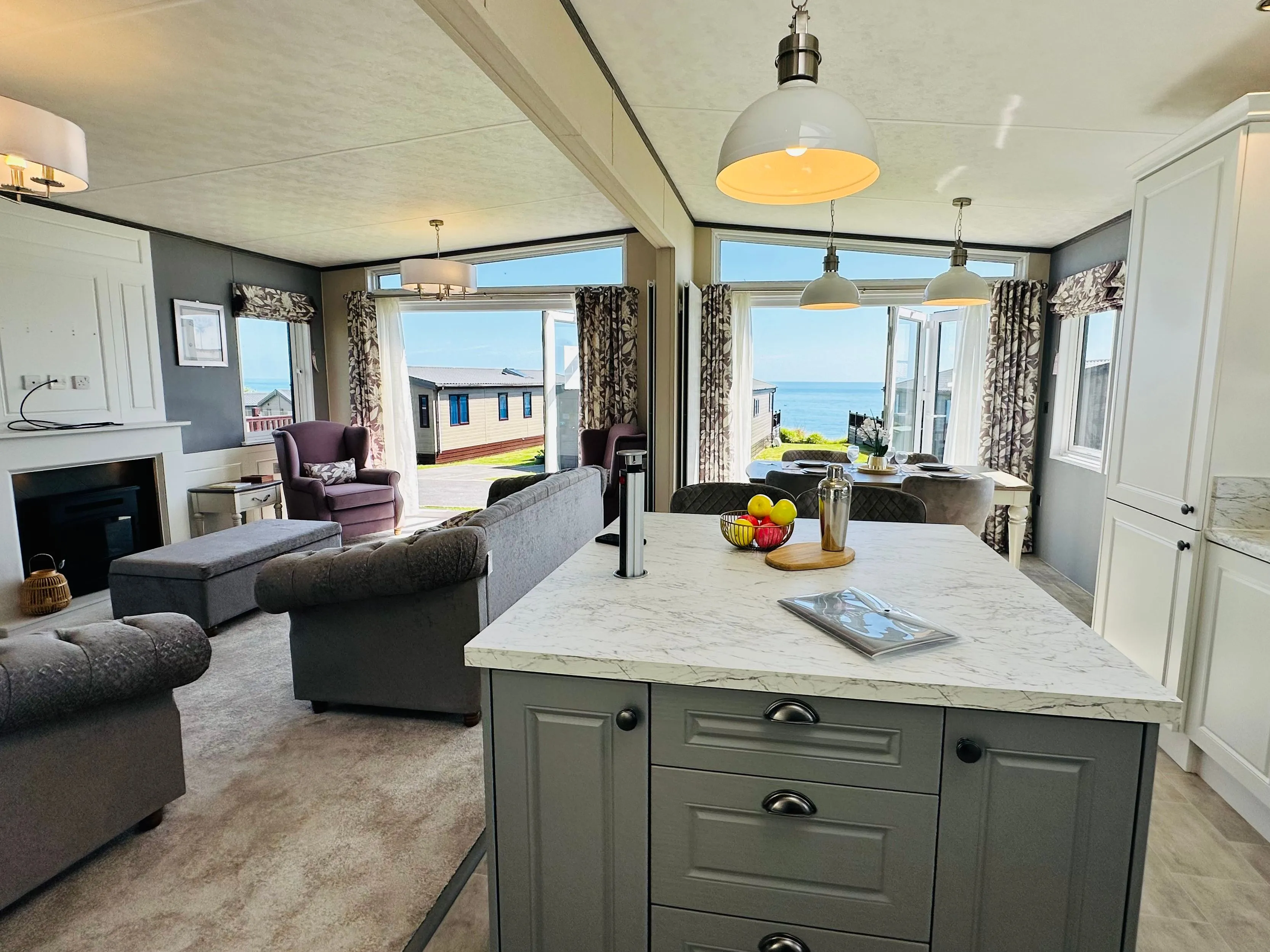 Modern open-plan holiday lodge interior with kitchen island, lounge area, dining space and sea view through large windows
