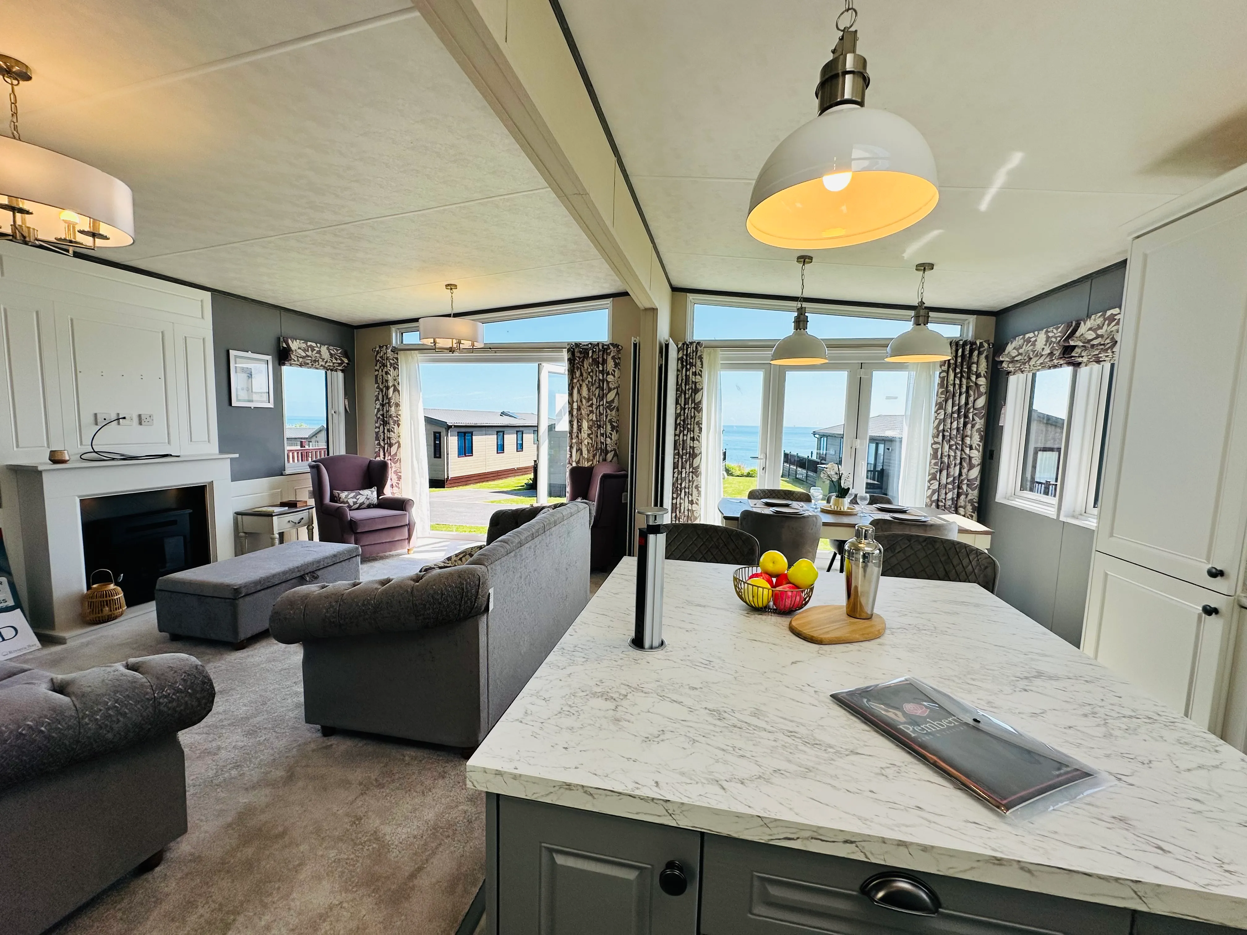 Modern open-plan holiday lodge interior with kitchen island, lounge, dining area and sea view through patio doors