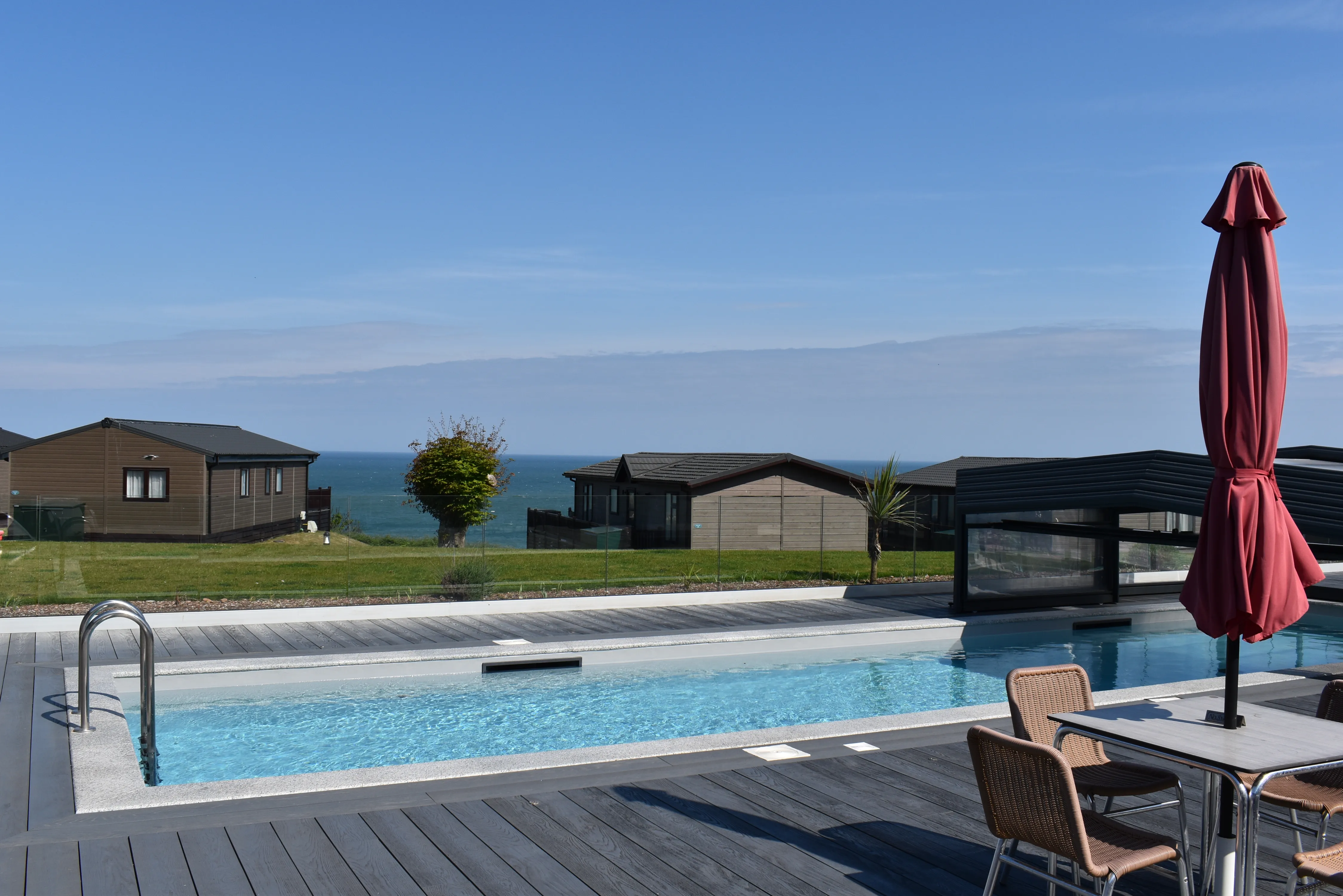 Outdoor swimming pool on a decking terrace overlooking coastal lodges and the sea under a clear blue sky