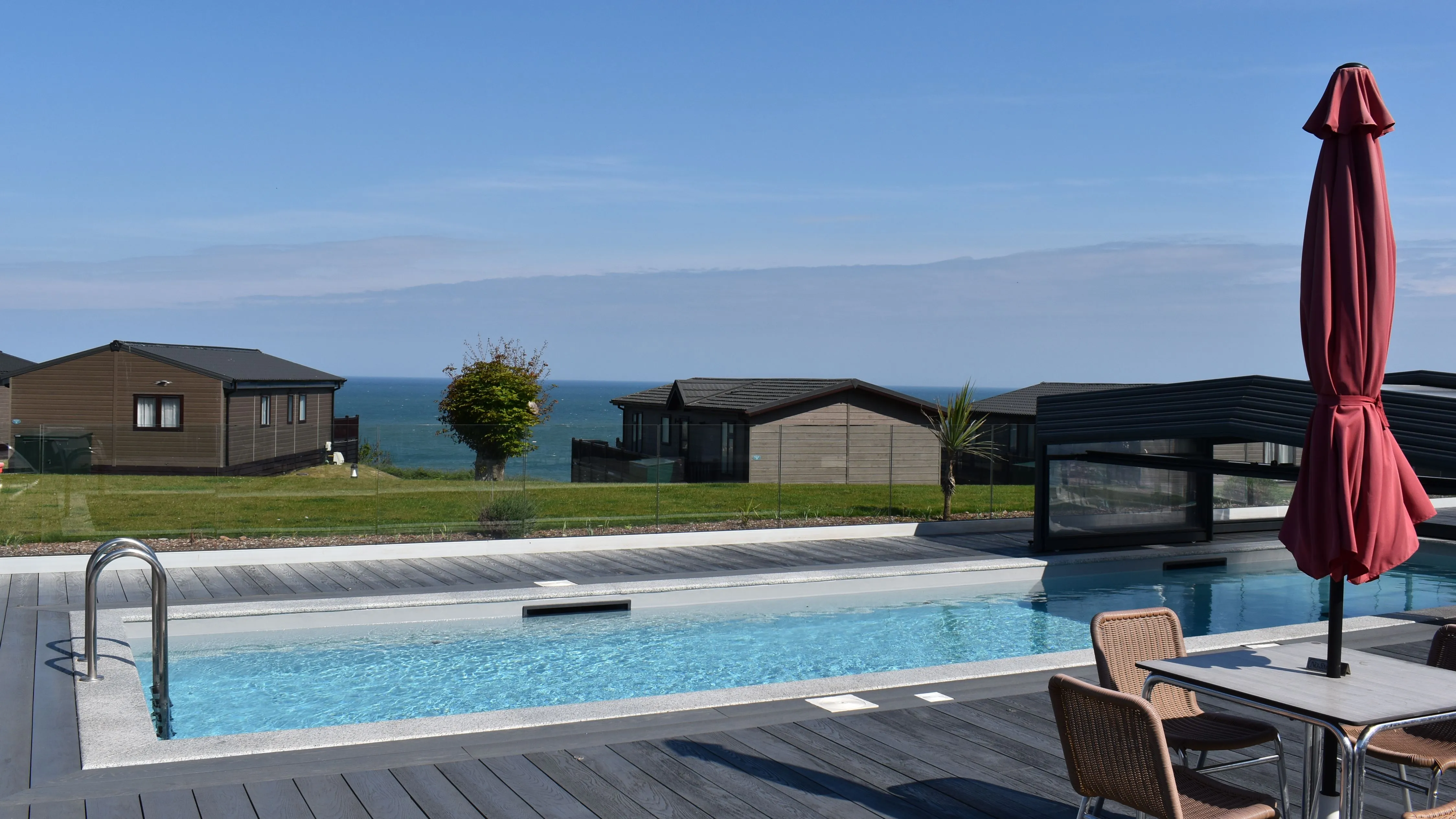 Outdoor swimming pool on a decking terrace overlooking coastal lodges and the sea under a clear blue sky