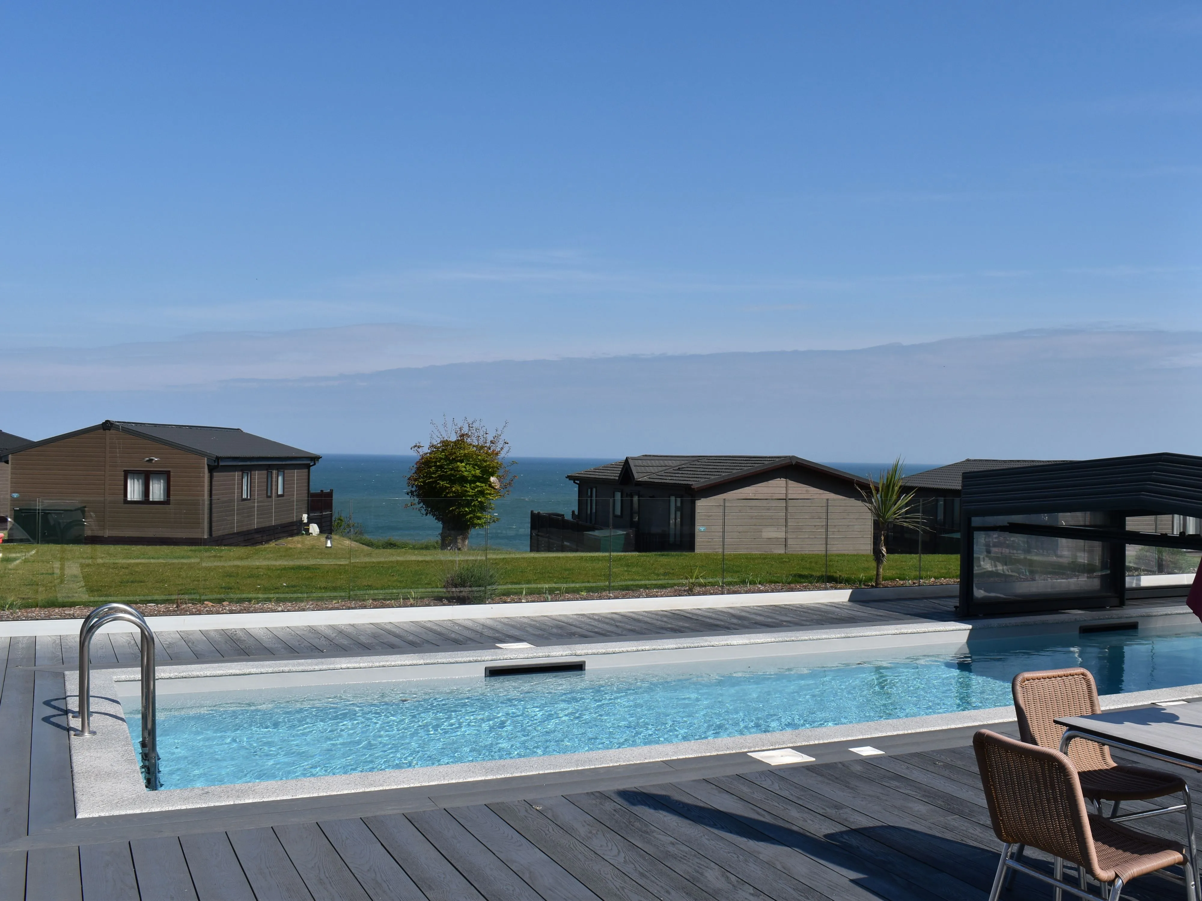 Outdoor swimming pool on a decking terrace overlooking coastal lodges and the sea under a clear blue sky