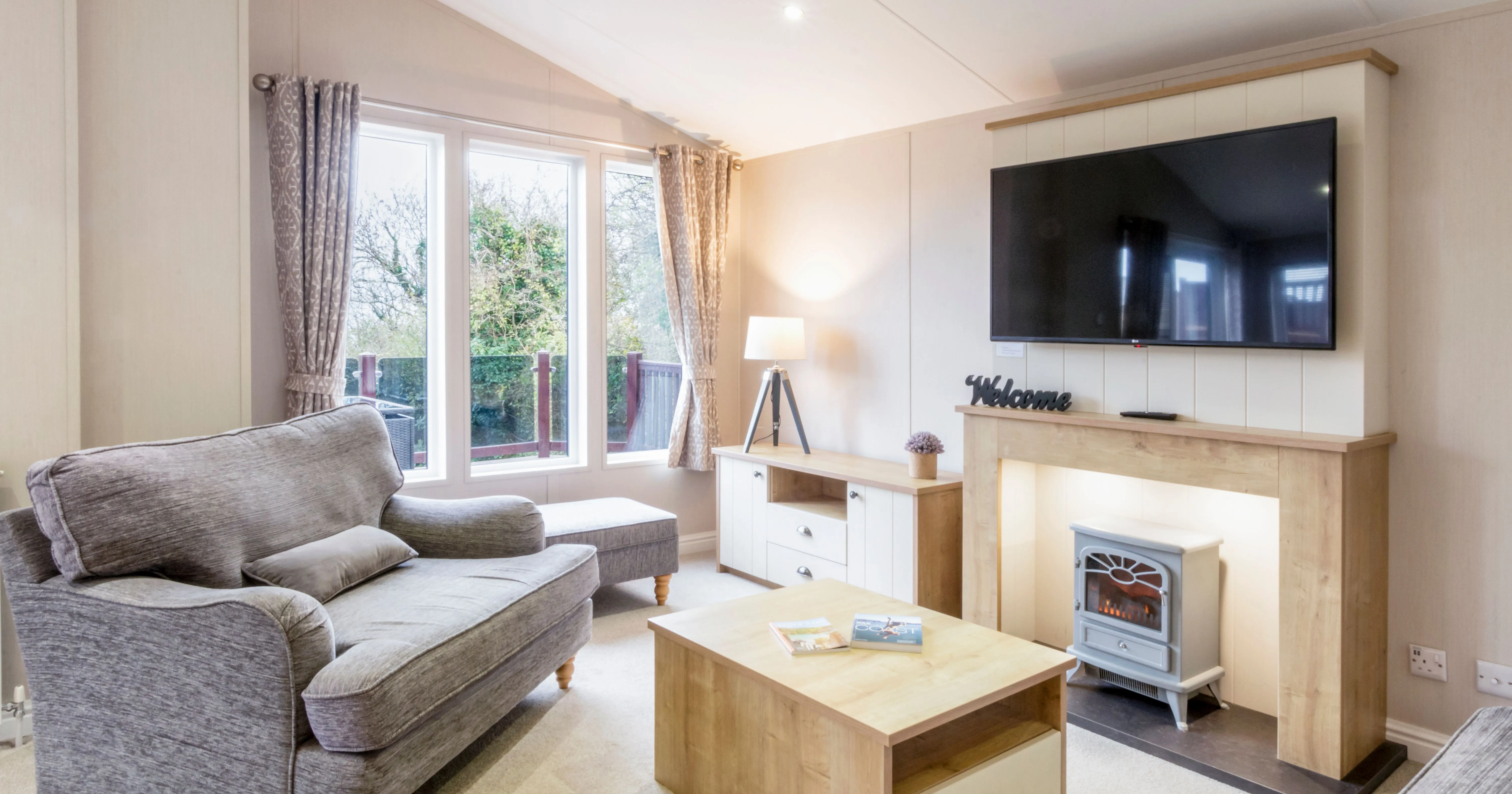 Bright modern living room with a grey sofa, large wall-mounted television, electric fireplace and tall windows with patterned curtains.