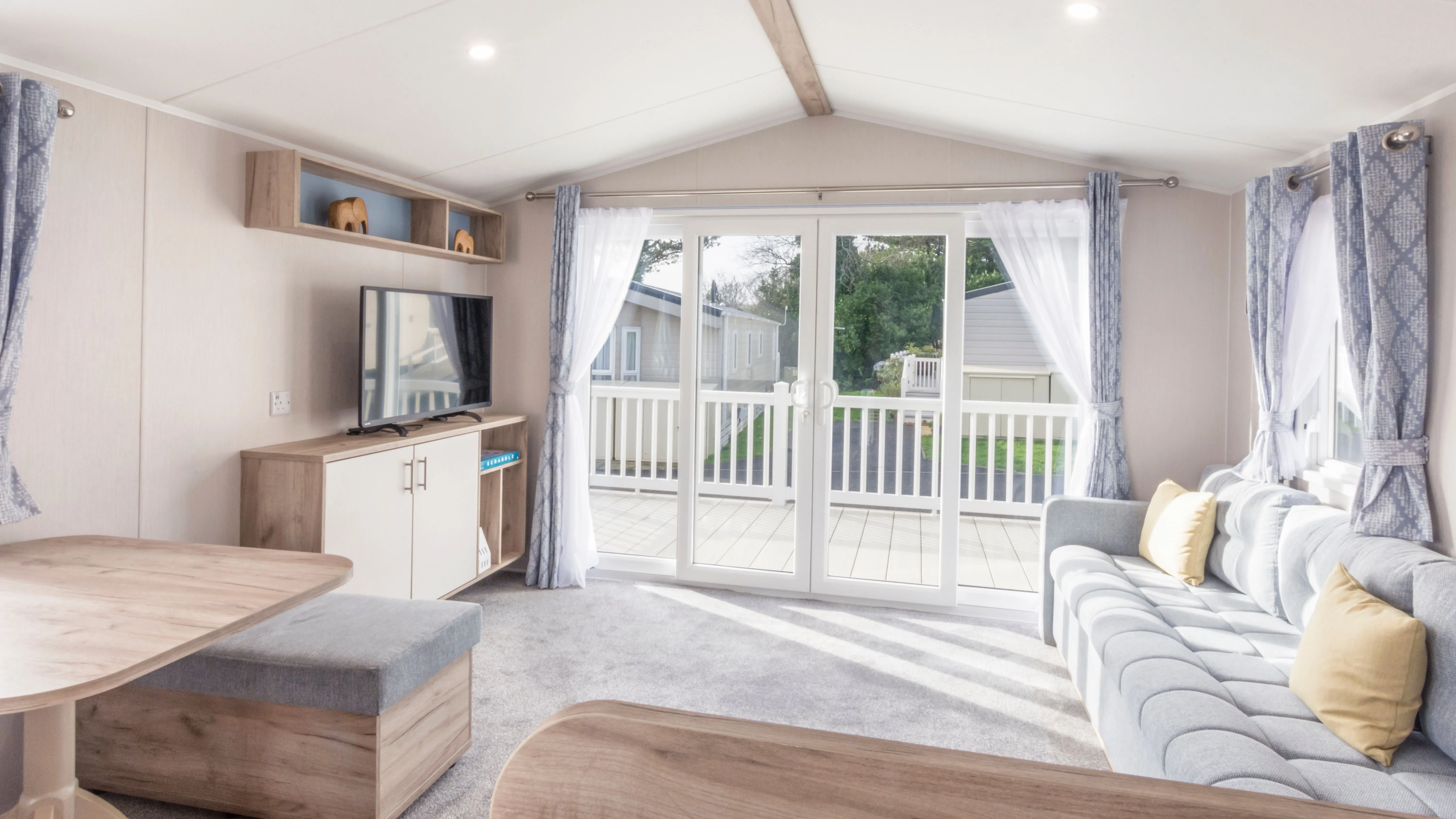 Bright modern caravan living room with grey sofa, TV unit, dining area and patio doors opening onto a veranda
