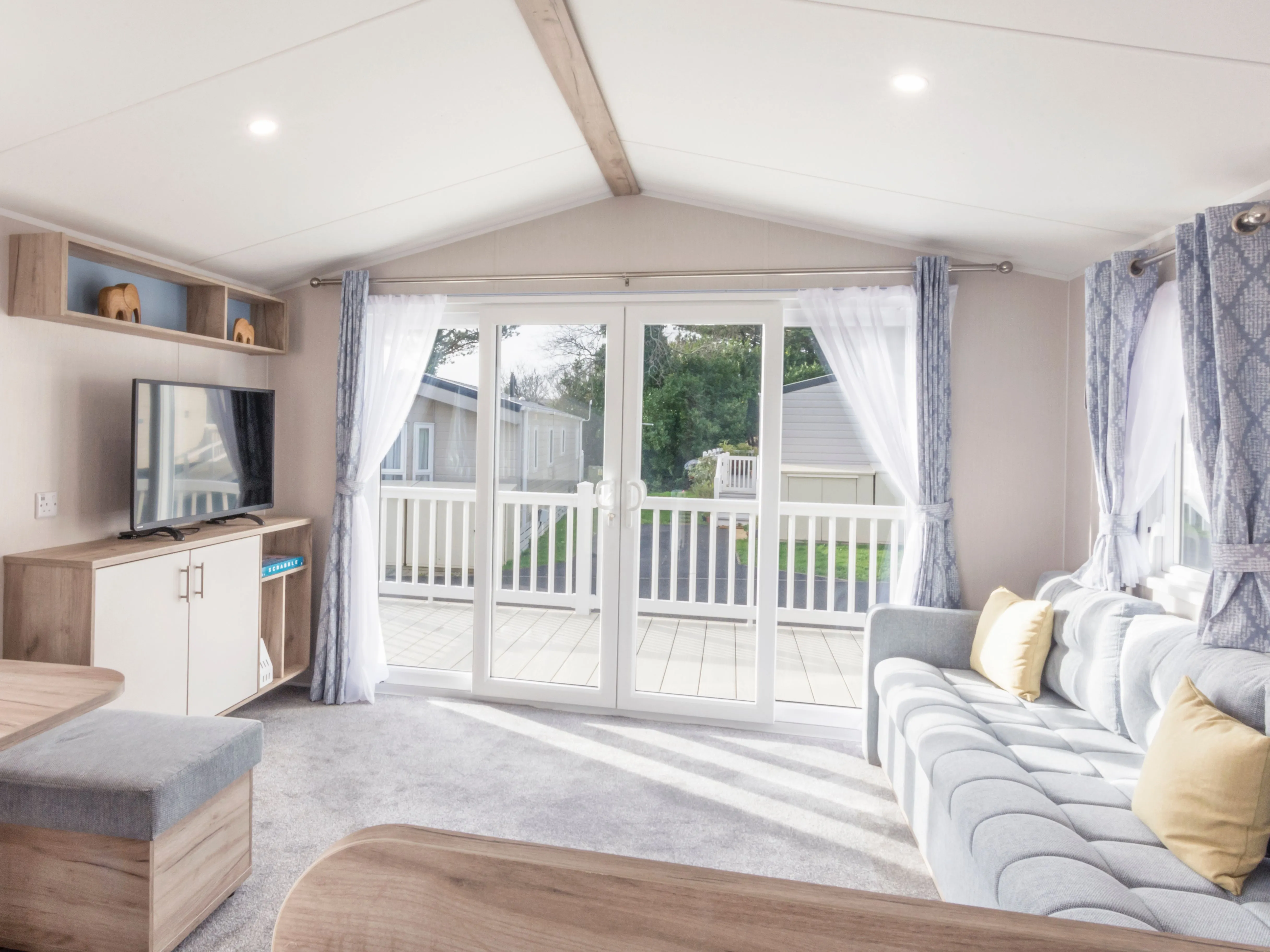 Bright modern caravan living room with grey sofa, TV unit, dining area and patio doors opening onto a veranda