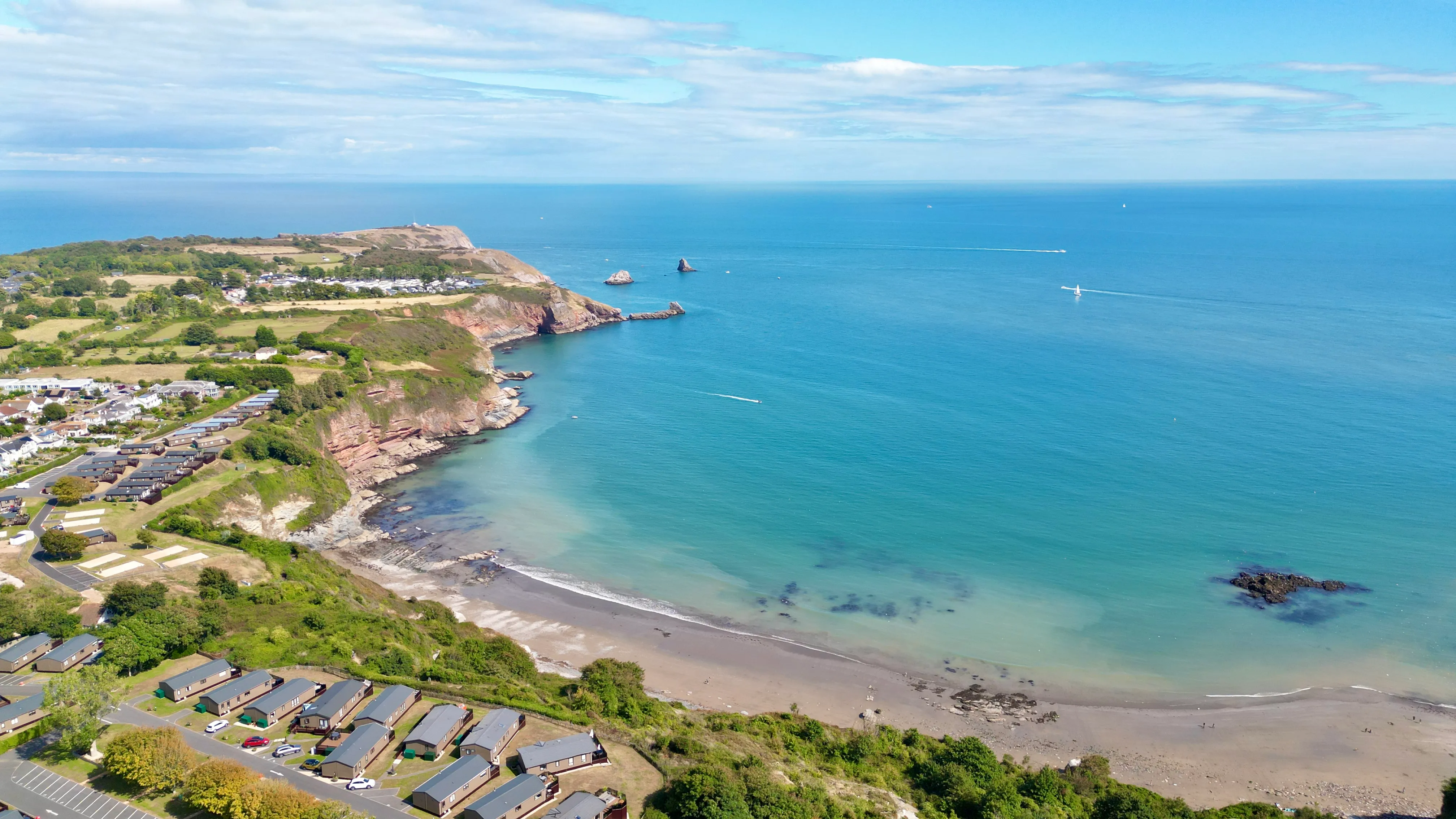 Drone view of Riviera Bay showing the sandy beach, turquoise sea, rocky cliffs and holiday park along the coastline.