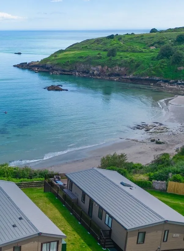 Drone view of Riviera Bay with holiday lodges overlooking a sandy beach, calm turquoise water and a green headland.