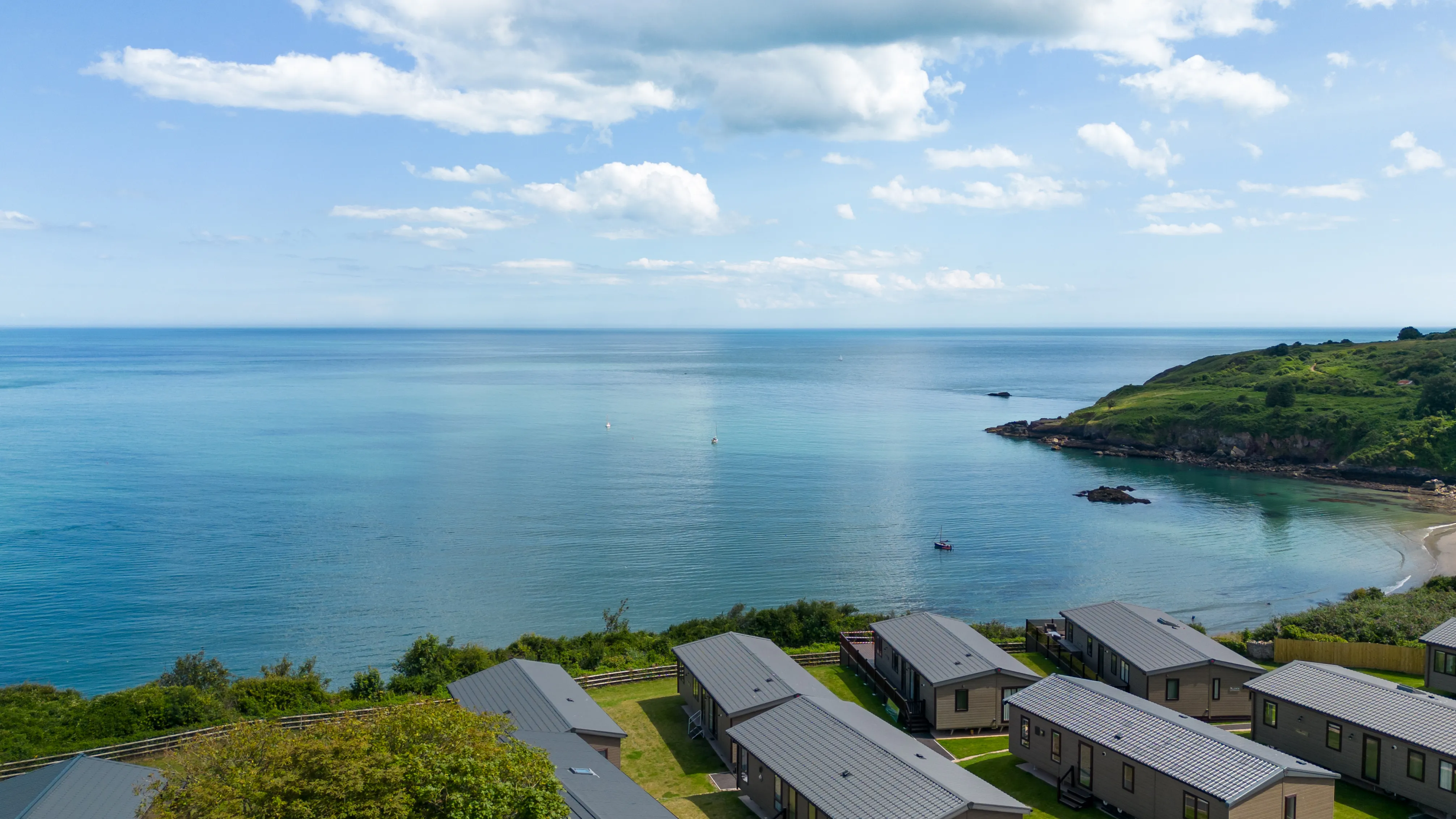 Drone view over Riviera Bay holiday park with coastal lodges, calm blue sea and a small sandy cove.