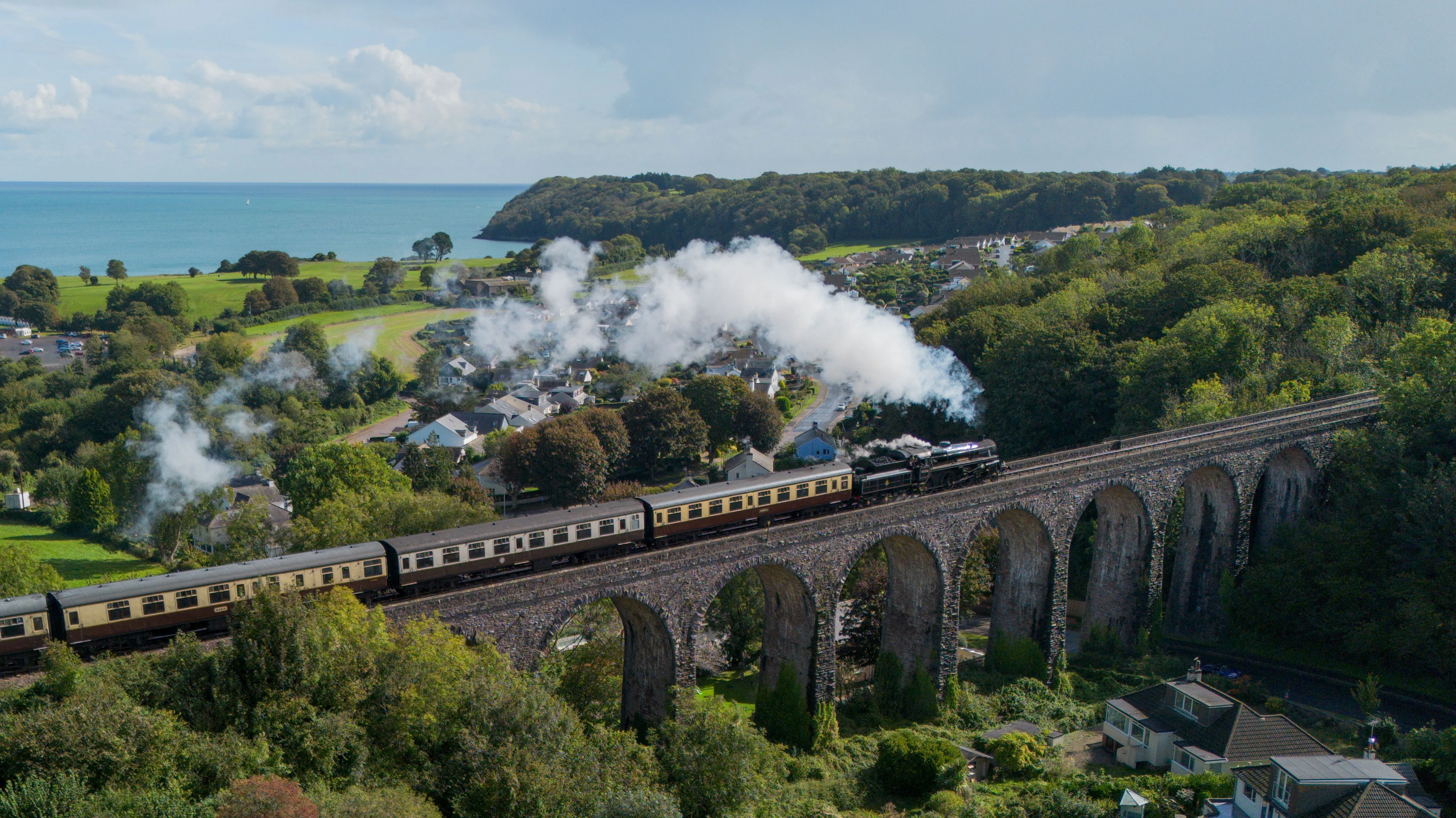 Steam train crossing a stone viaduct above a coastal village with sea and woodland in the background