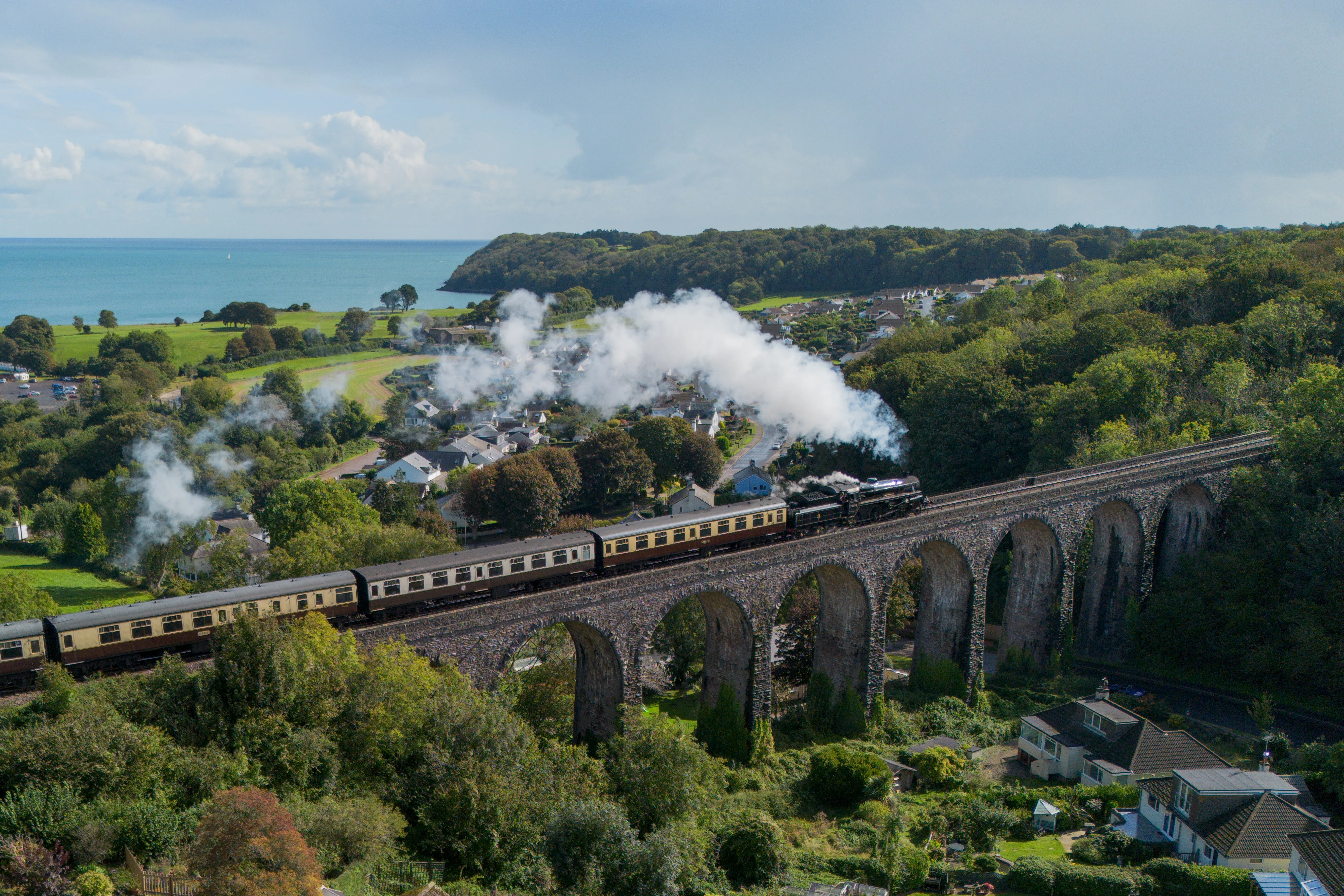 Steam train crossing a stone viaduct above a coastal village with sea and woodland in the background