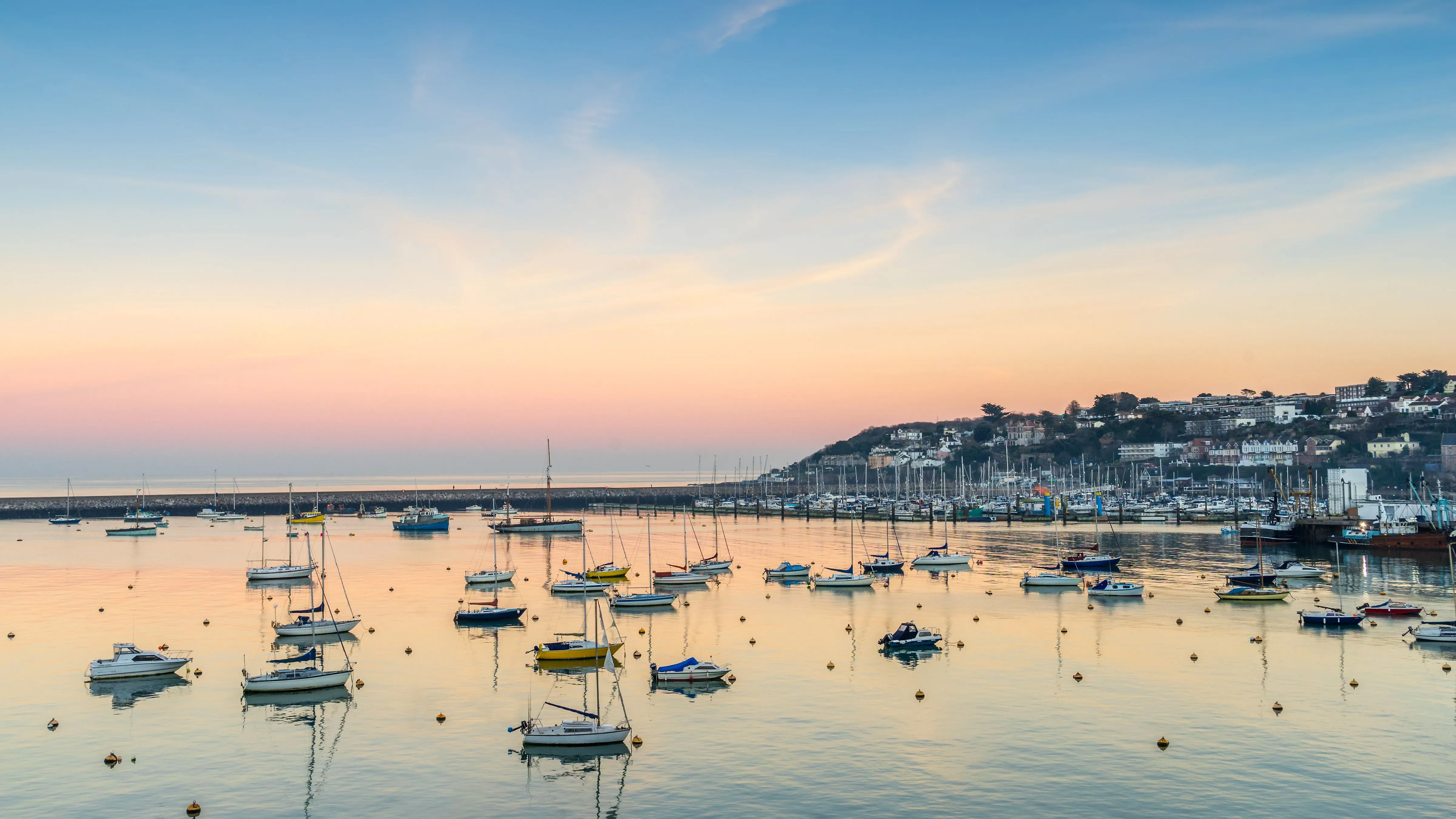 Sailing boats moored in a calm harbour at sunset, with pastel skies and a coastal town on the hillside