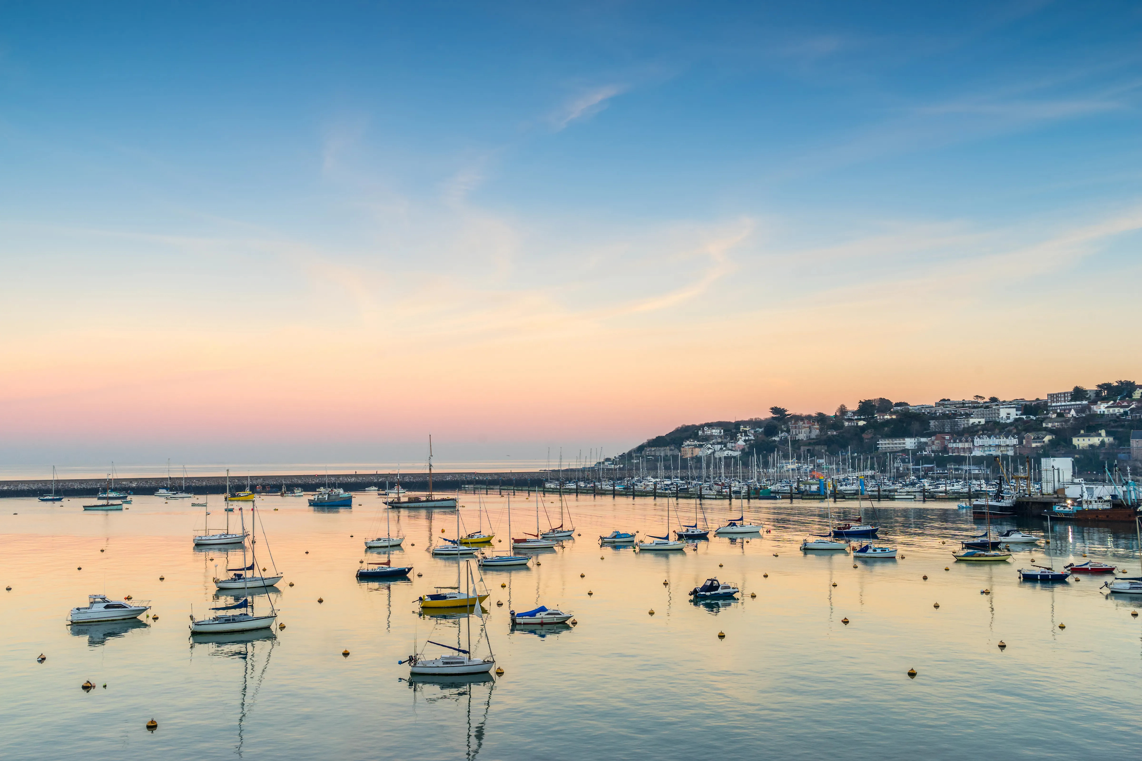 Sailing boats moored in a calm harbour at sunset, with pastel skies and a coastal town on the hillside