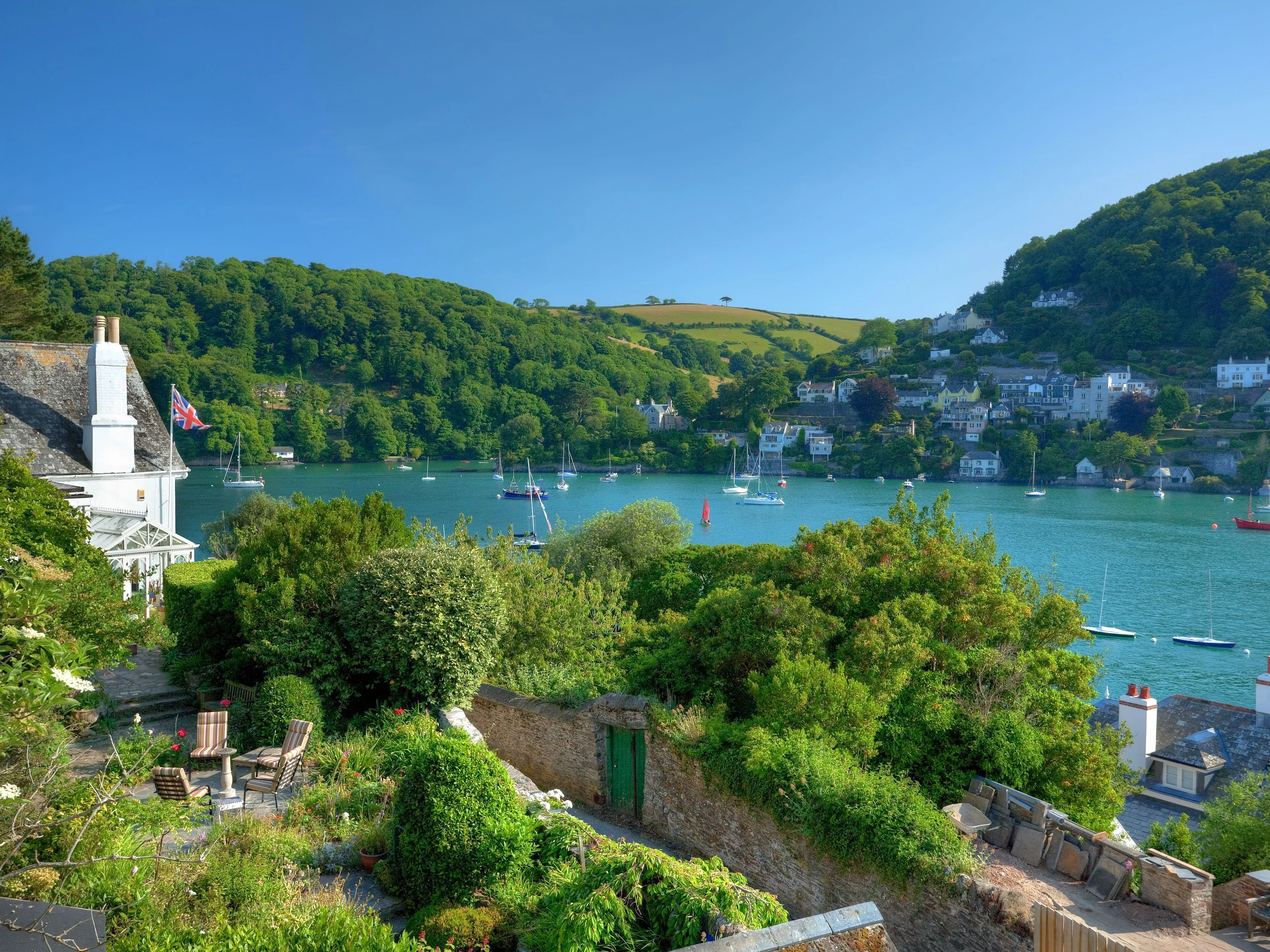 View across a tranquil estuary with sailing boats, hillside cottages and a lush garden beside a white house flying a Union flag.