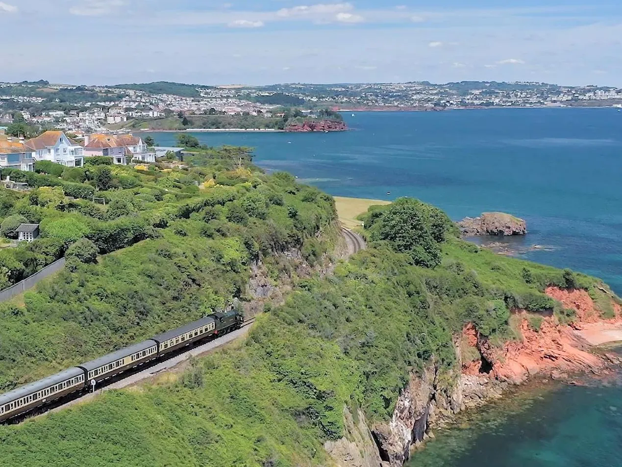 Train travelling along a coastal cliff railway beside blue sea and hillside houses