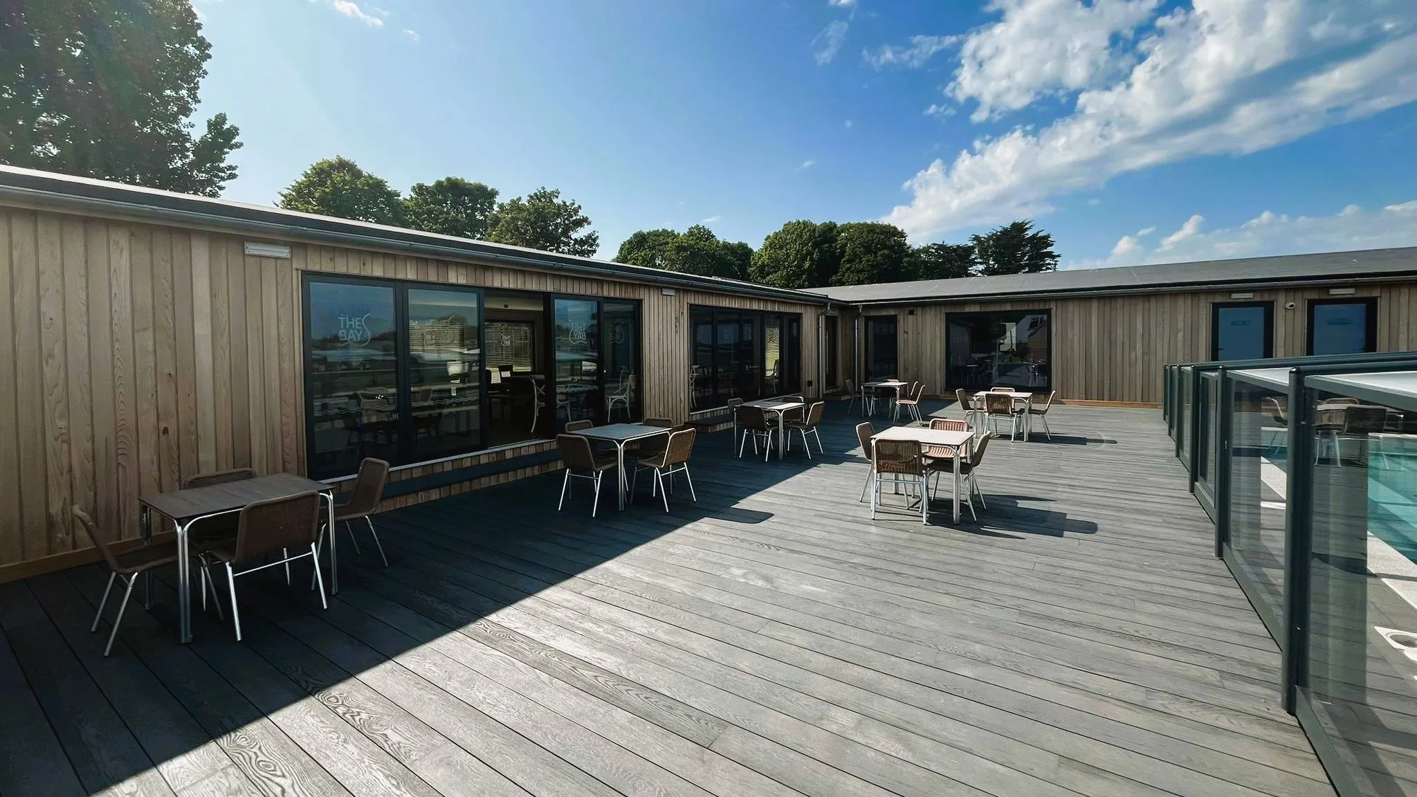 Spacious outdoor decking area with tables and chairs beside a modern timber-clad building under a bright blue sky.