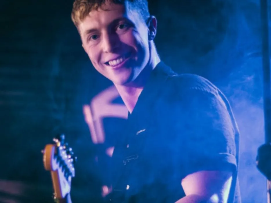 Jamie smiling on stage with an electric guitar under blue and purple lighting