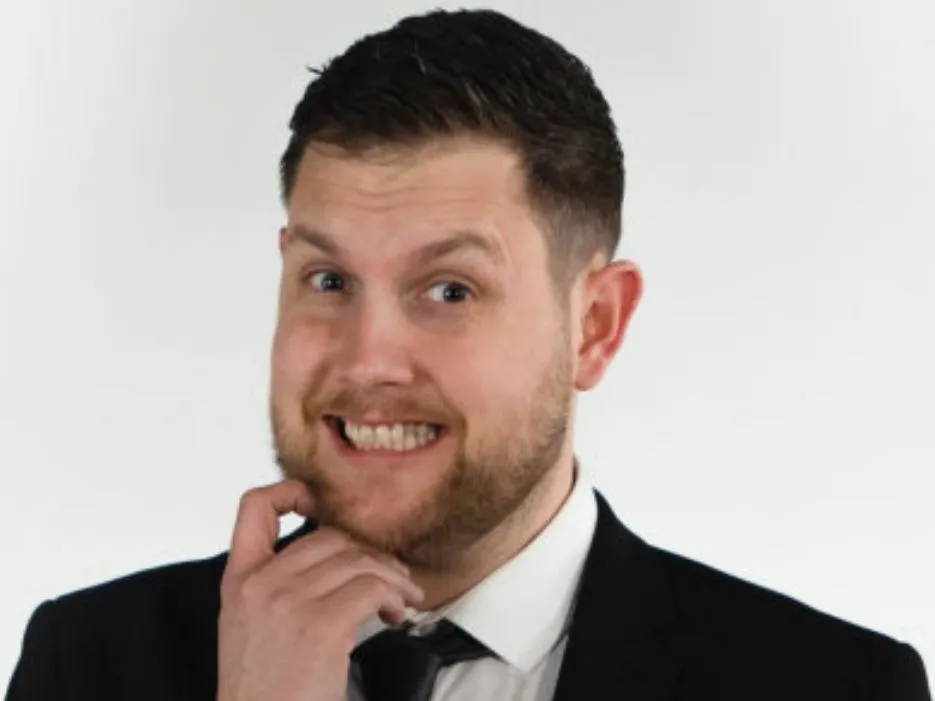 Joe Stone smiling in a suit with his hand resting on his chin against a plain white background