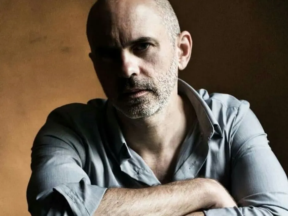 Portrait of Ed Scott leaning forward with folded arms, wearing a light grey shirt against a warm brown background
