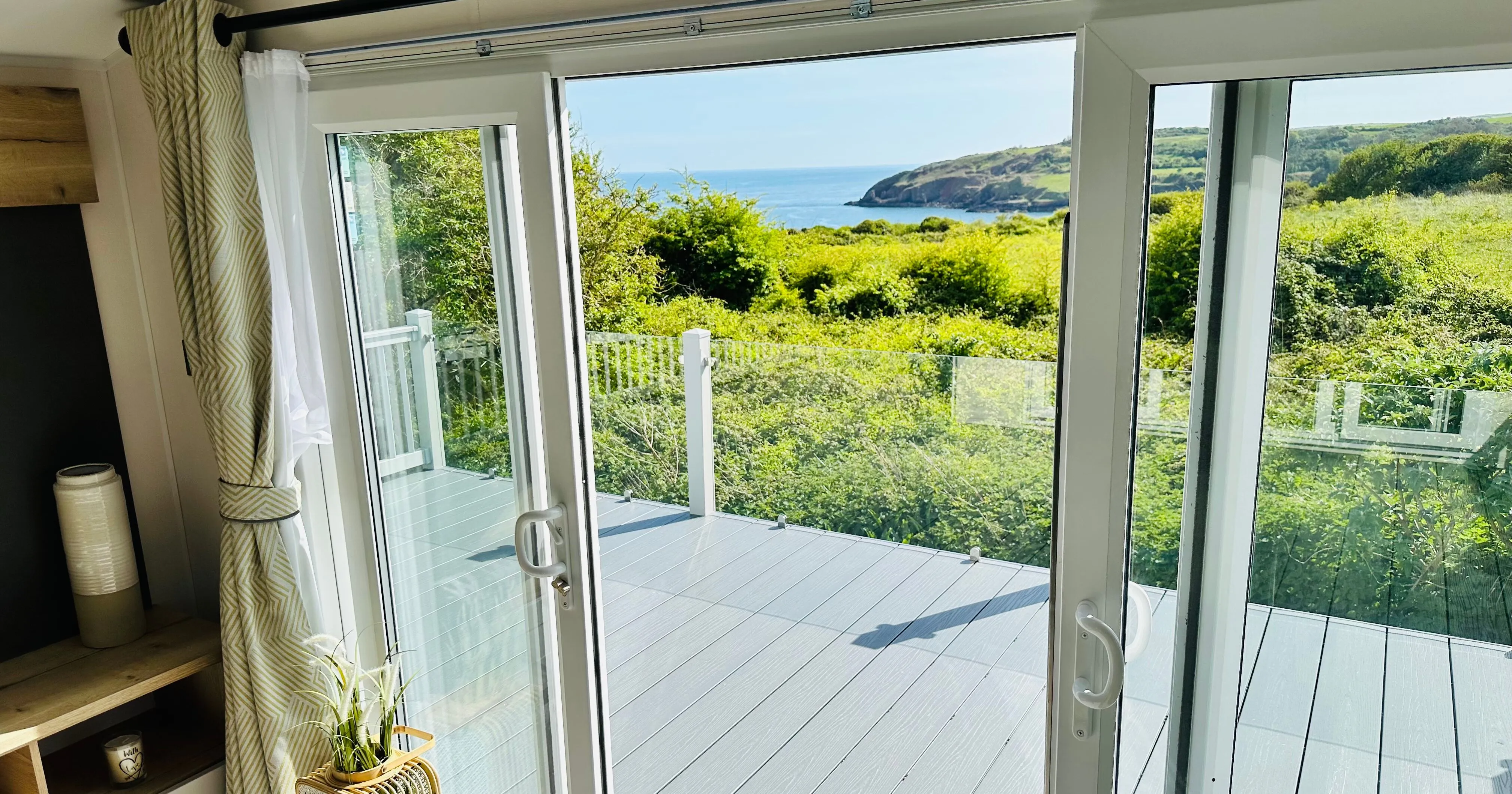 Open patio doors leading to a balcony with glass balustrades and a sea view beyond green coastal countryside