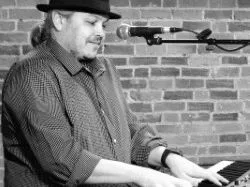 Performer in a hat playing an electronic keyboard beside a microphone against a brick wall