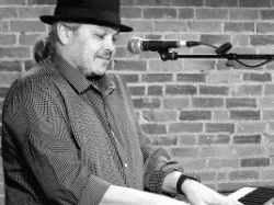 Performer in a hat playing an electronic keyboard beside a microphone against a brick wall