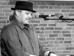 Performer in a hat playing an electronic keyboard beside a microphone against a brick wall