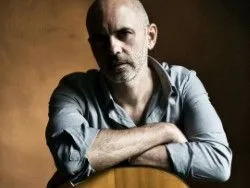 Portrait of a bald bearded man leaning on an acoustic guitar against a dark background