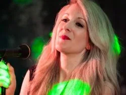 Blonde female singer performing into a microphone under green stage lighting