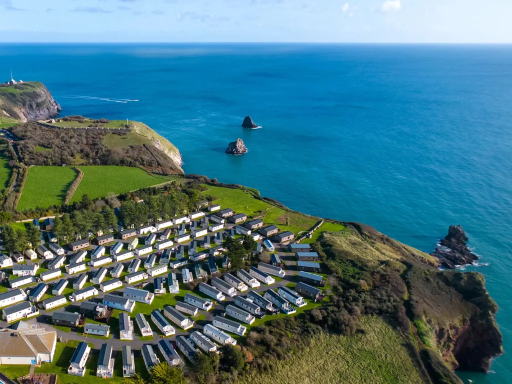 Aerial view of a coastal holiday park with static caravans on green clifftops overlooking a bright blue sea and rocky outcrops.