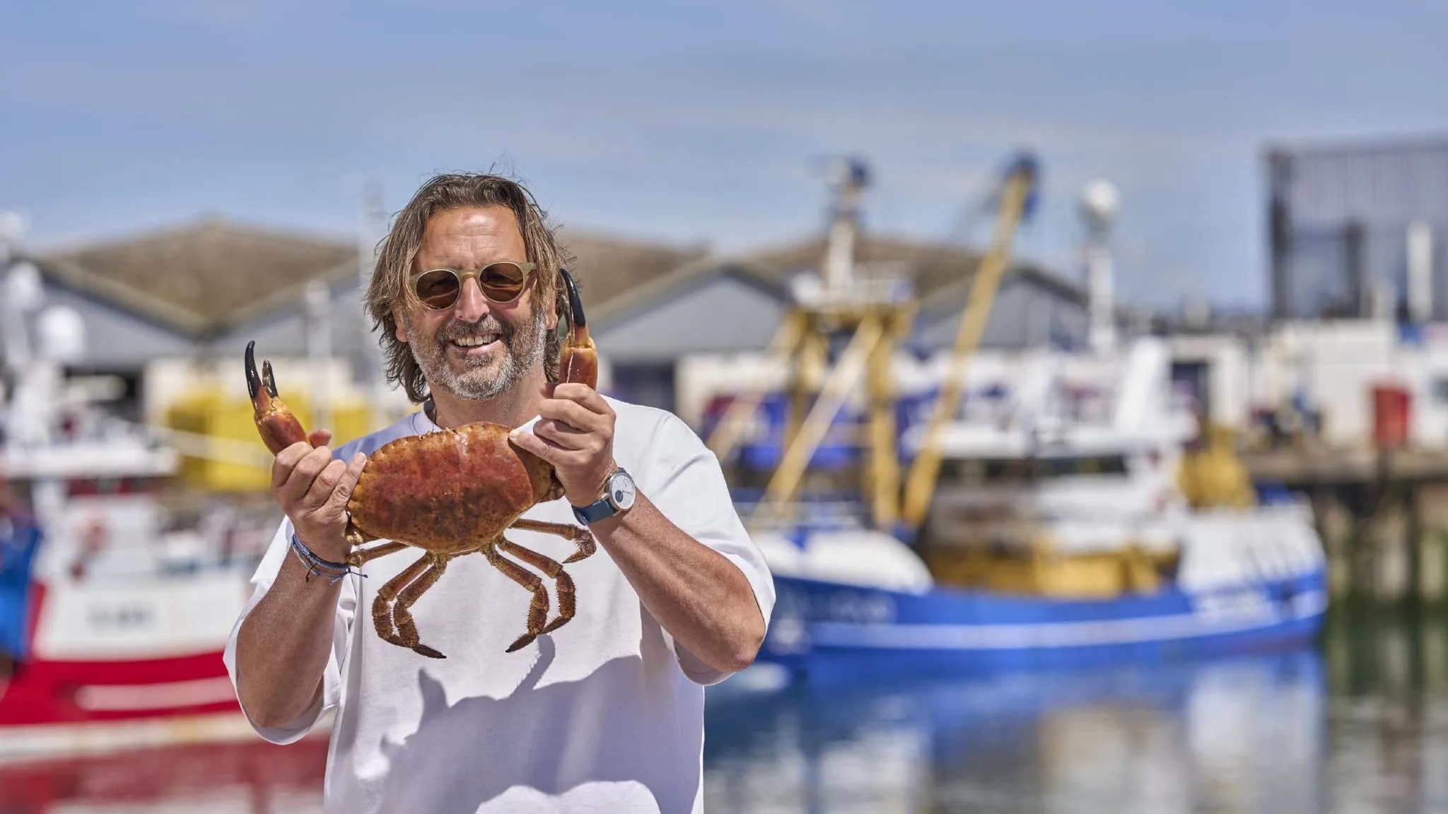 Man holding a large crab at a harbour with fishing boats in the background