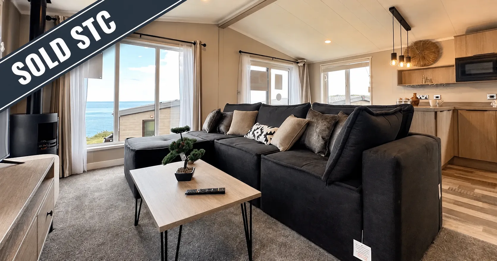 Modern lodge living room with a large dark sectional sofa, light wood coffee table, sea view through tall windows, and an open-plan kitchen, marked Sold STC.