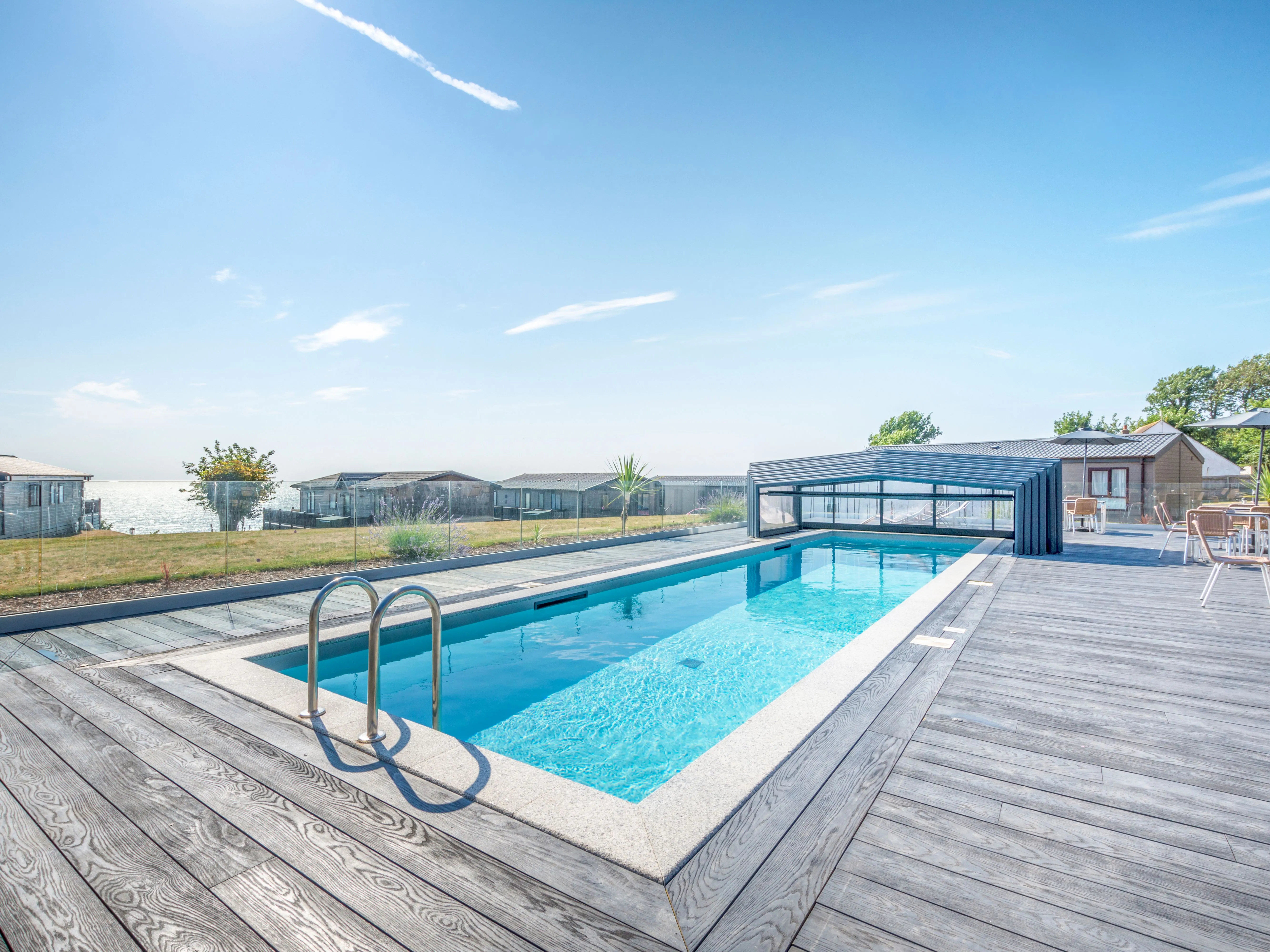 Outdoor swimming pool with a timber deck, retractable cover and sea view beside holiday lodges.
