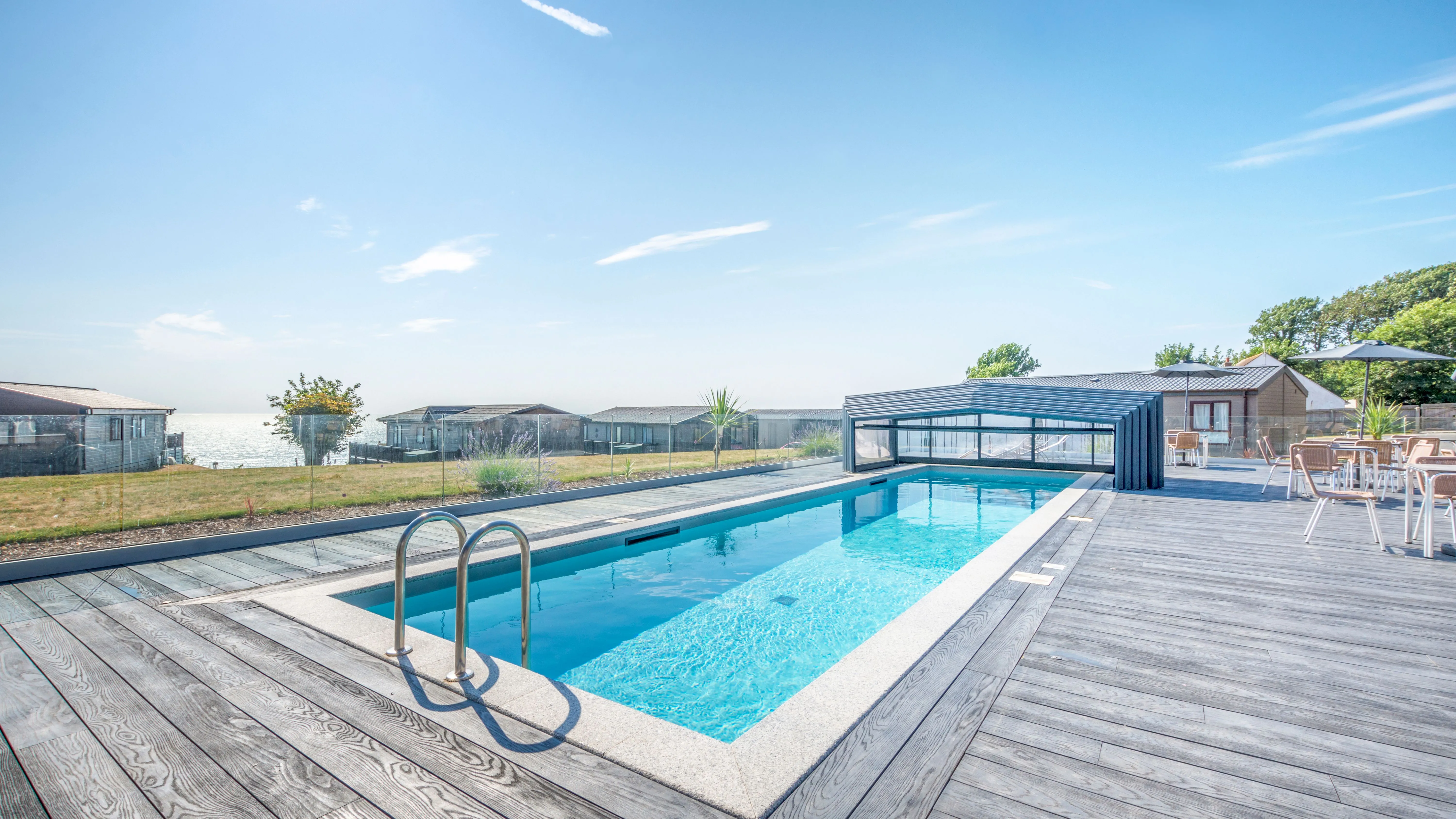 Outdoor swimming pool with a timber deck, retractable cover and sea view beside holiday lodges.