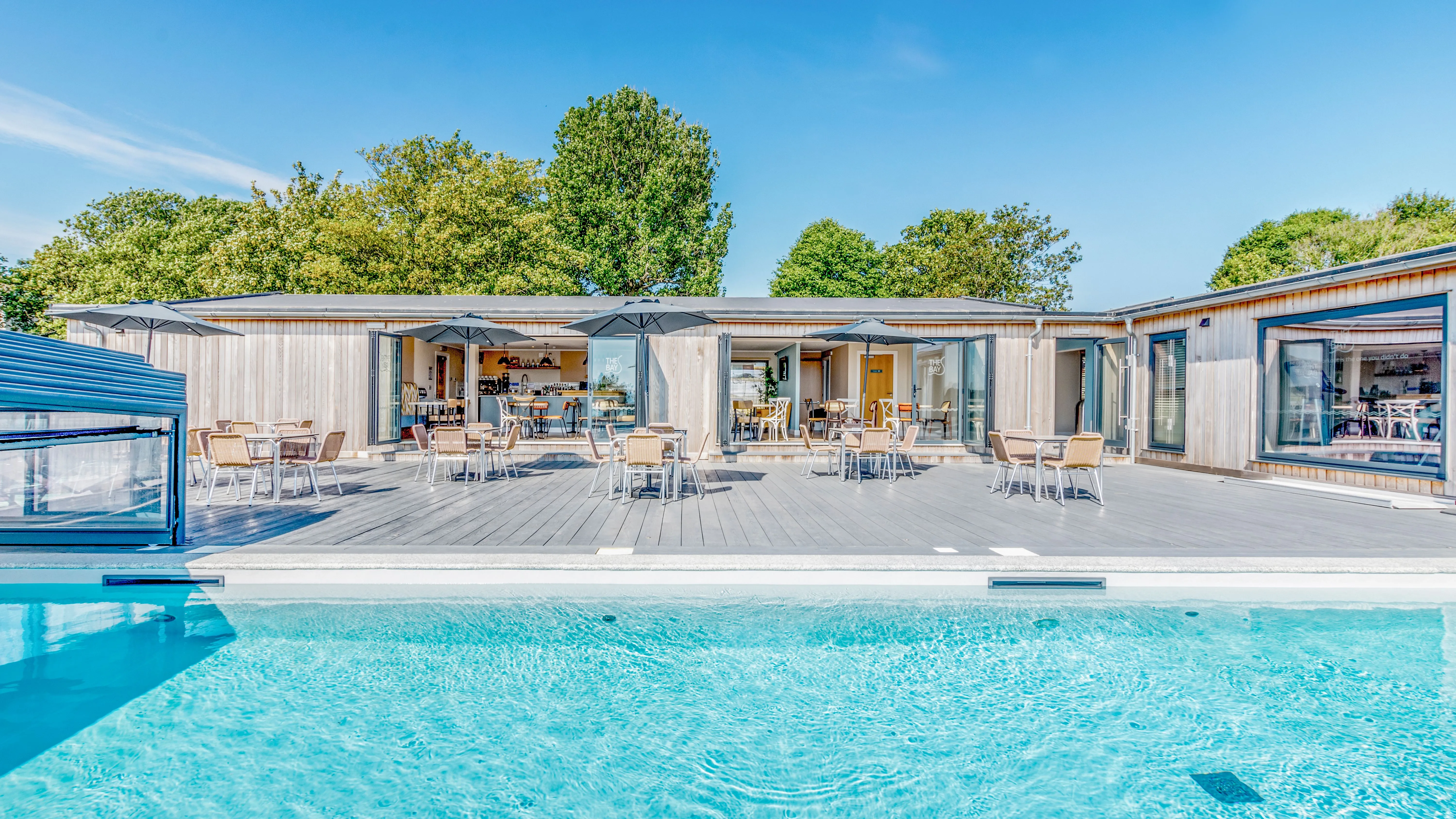 Outdoor swimming pool beside a modern wooden café terrace with tables, chairs and parasols under a clear blue sky.