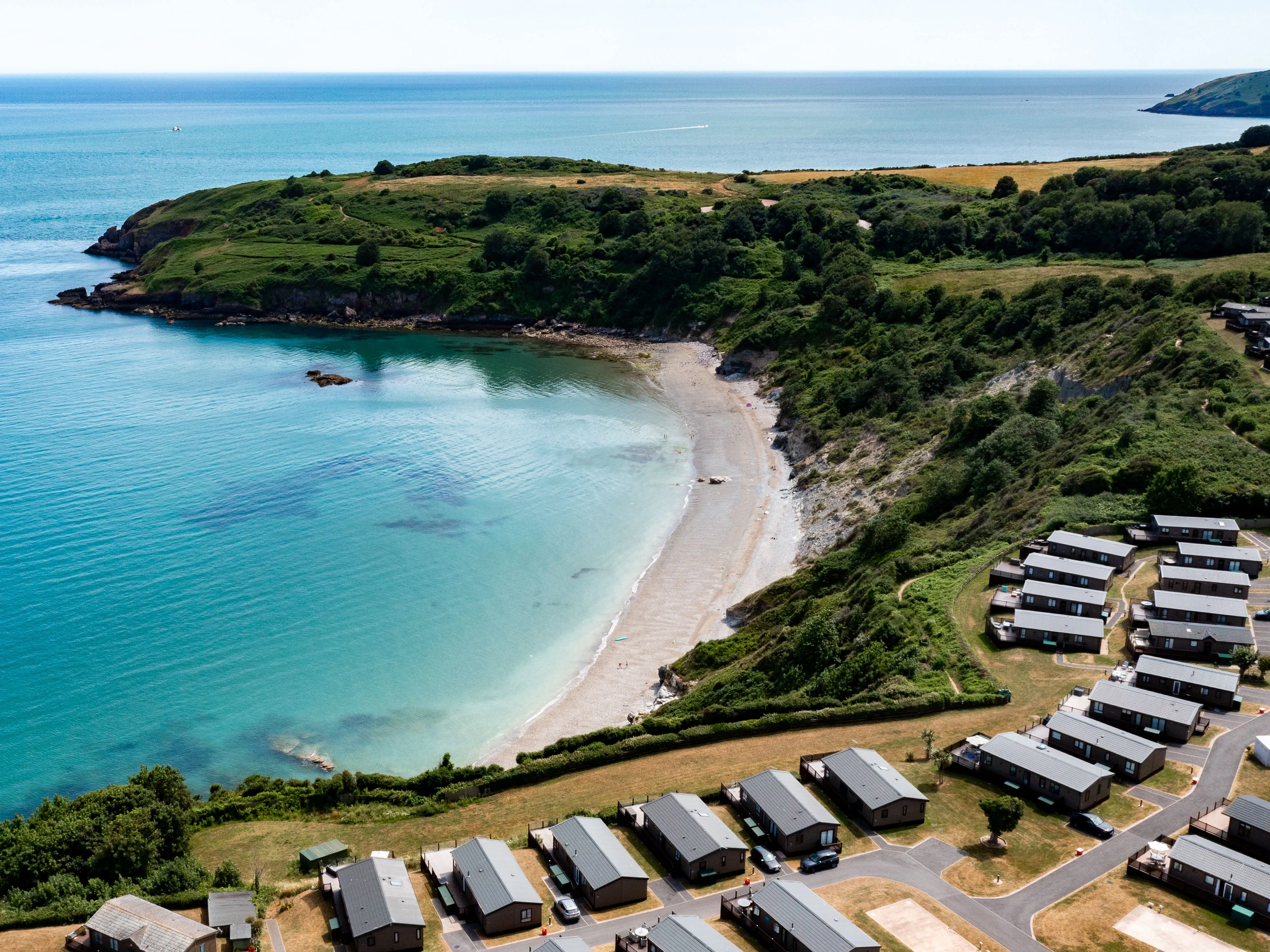 Aerial view of a secluded crescent beach with turquoise water, green coastal cliffs and holiday lodges.