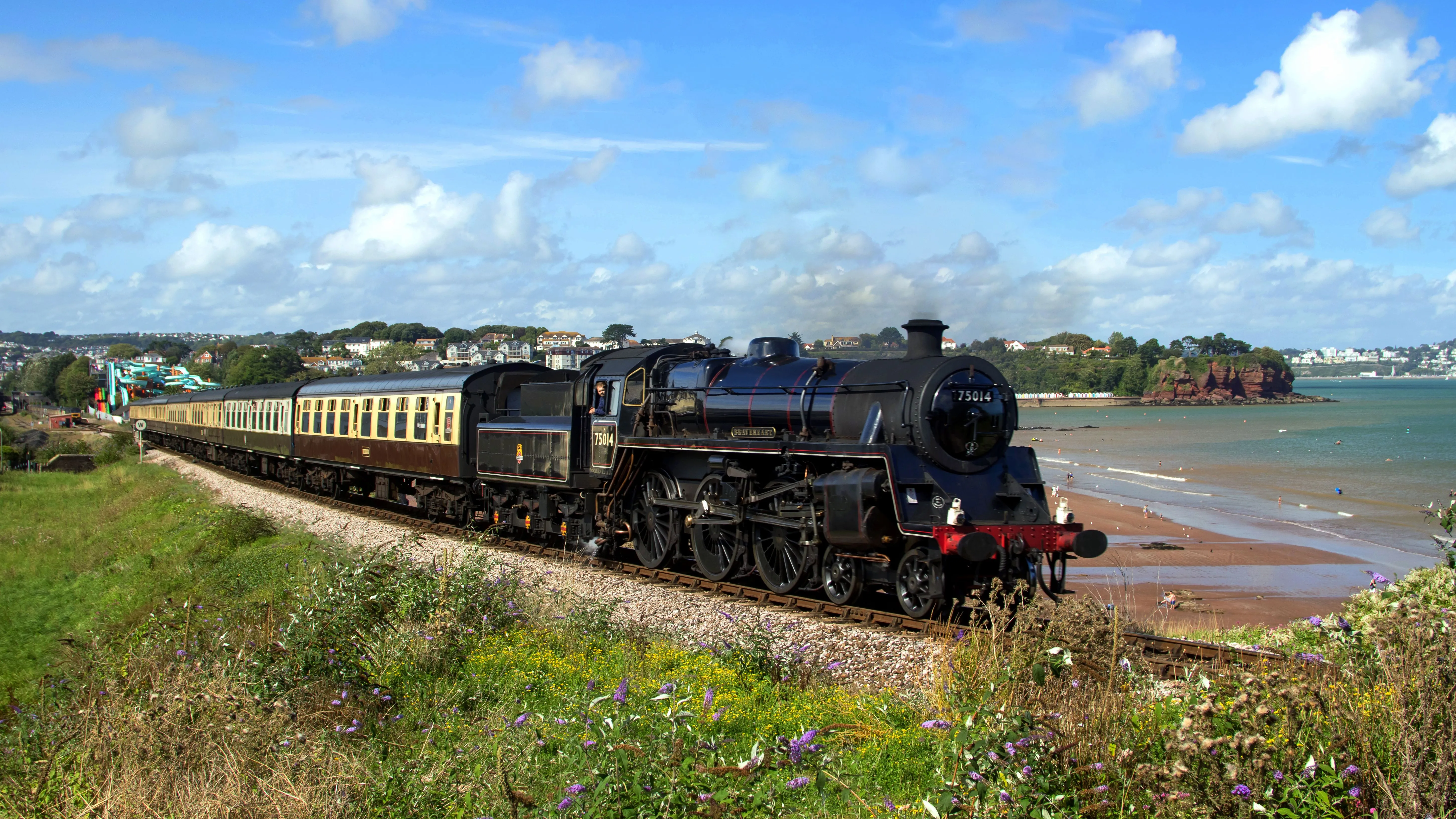 Steam locomotive Braveheart, number 75014, hauling vintage carriages along the coastal railway at Goodrington beside a sandy beach.